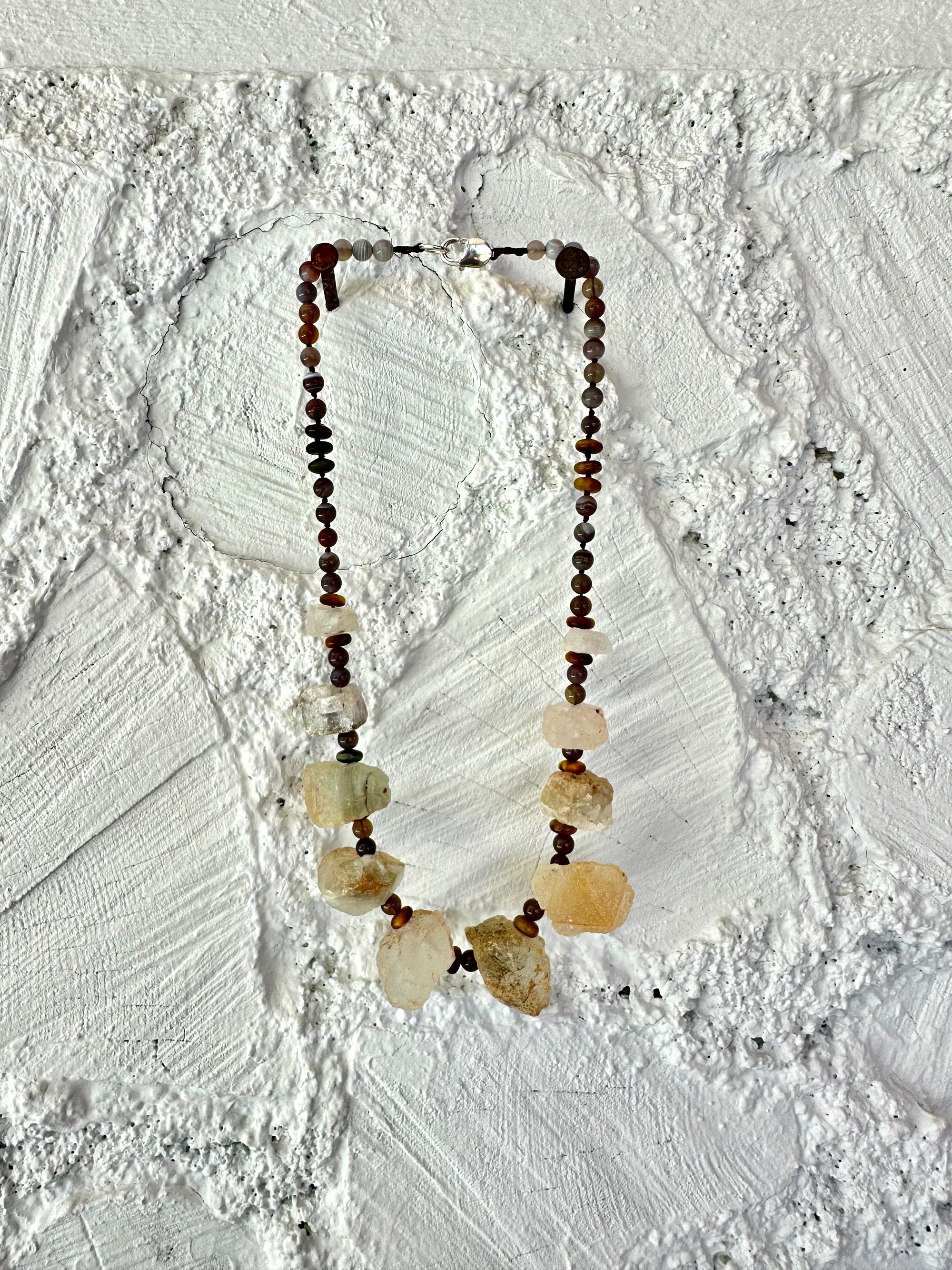 10 light green nuggets of fluorite are joined by dark, gray and brown beads of egg and tiger eye in a necklace, hanging on a white painted cordwood wall