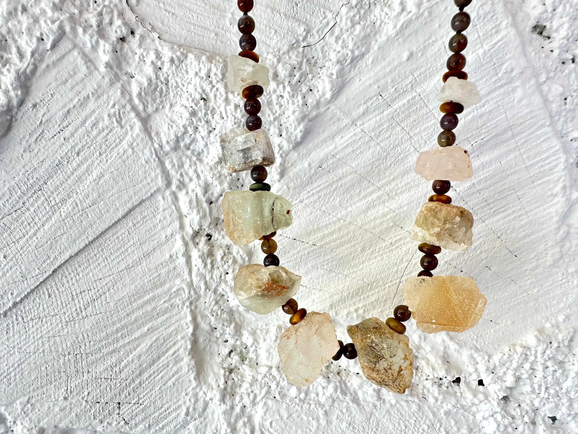 10 light green nuggets of fluorite are joined by dark, gray and brown beads of egg and tiger eye in a necklace, hanging on a white painted cordwood wall