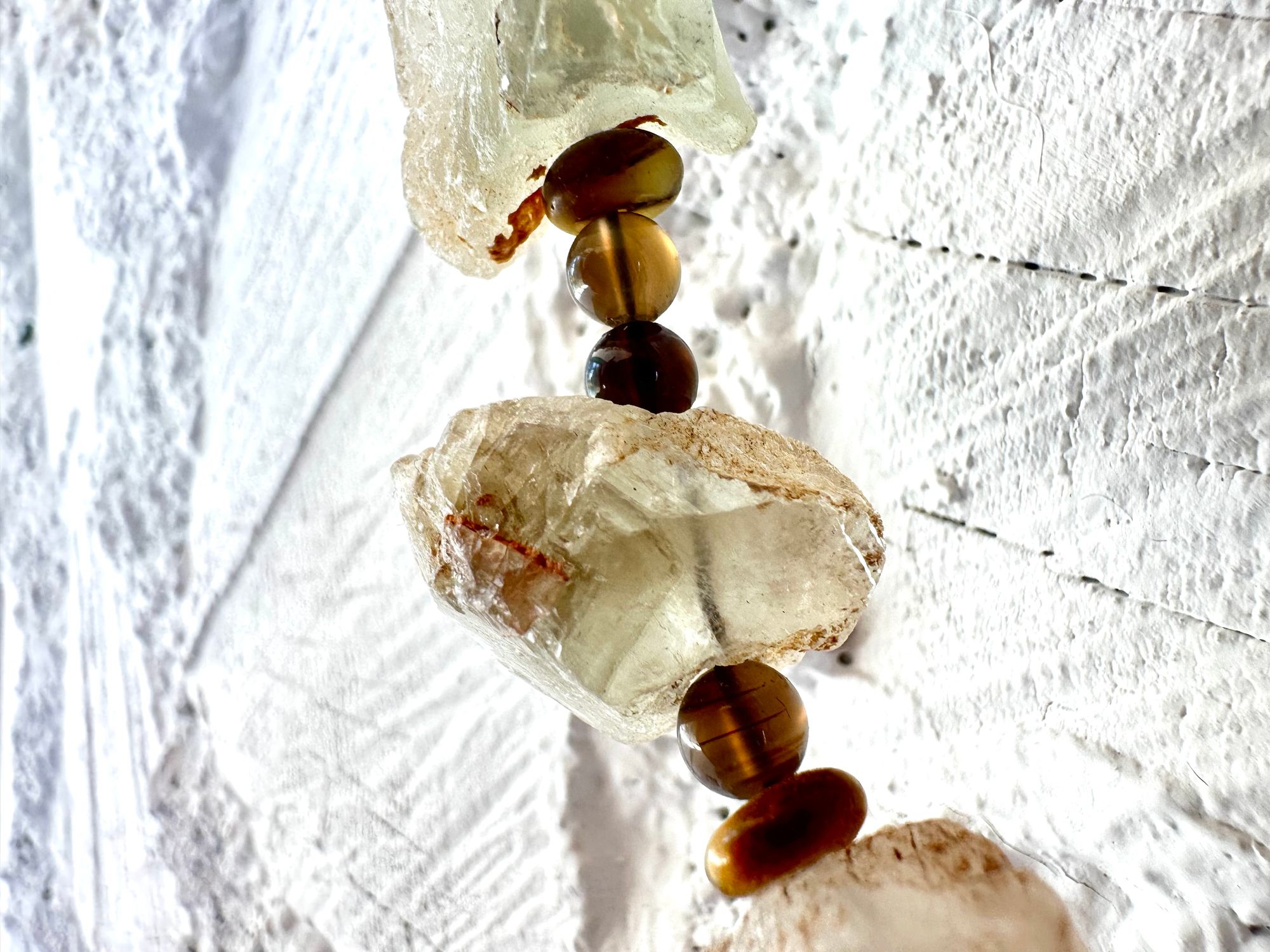 10 light green nuggets of fluorite are joined by dark, gray and brown beads of egg and tiger eye in a necklace, hanging on a white painted cordwood wall