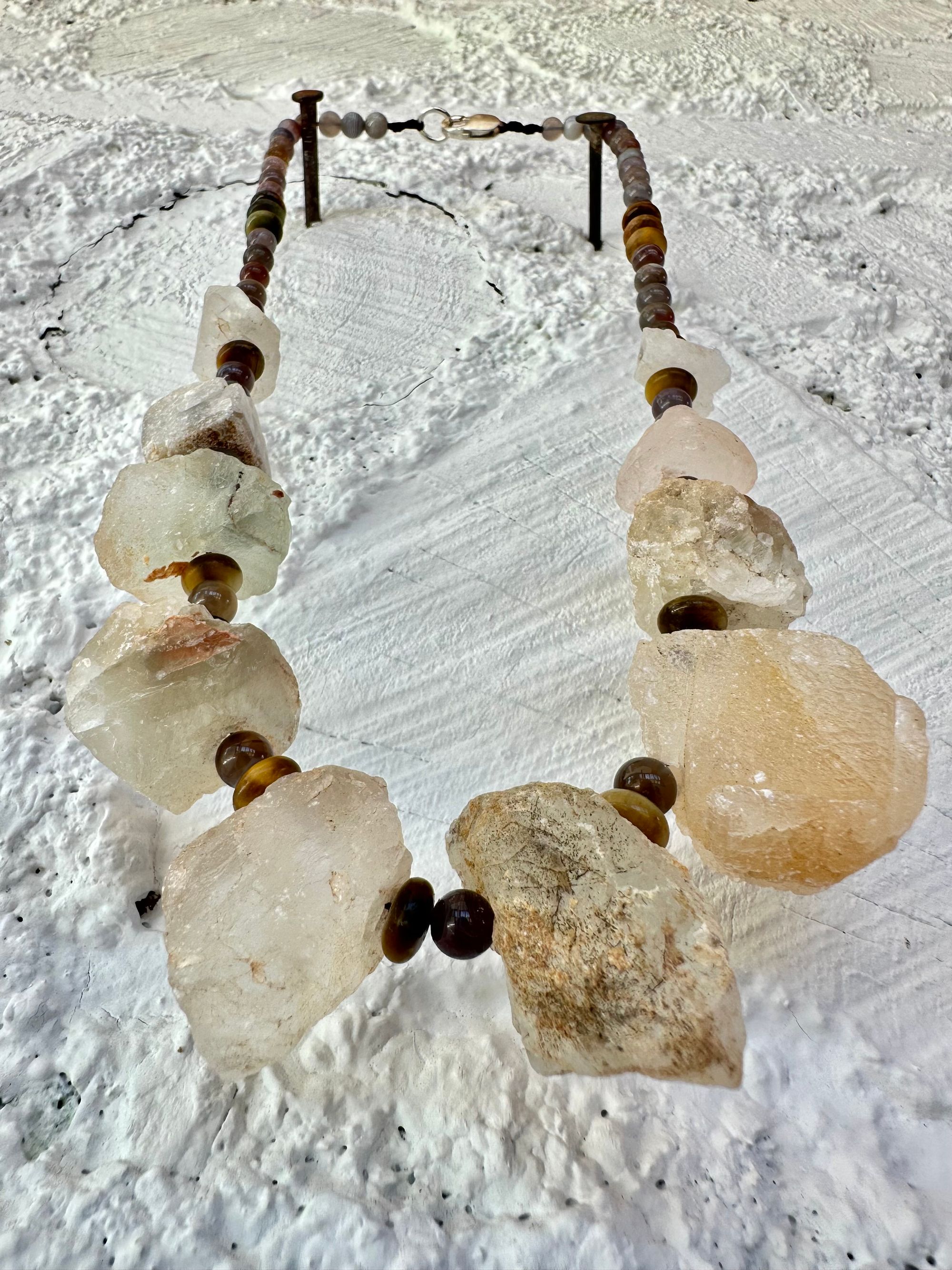 10 light green nuggets of fluorite are joined by dark, gray and brown beads of egg and tiger eye in a necklace, hanging on a white painted cordwood wall