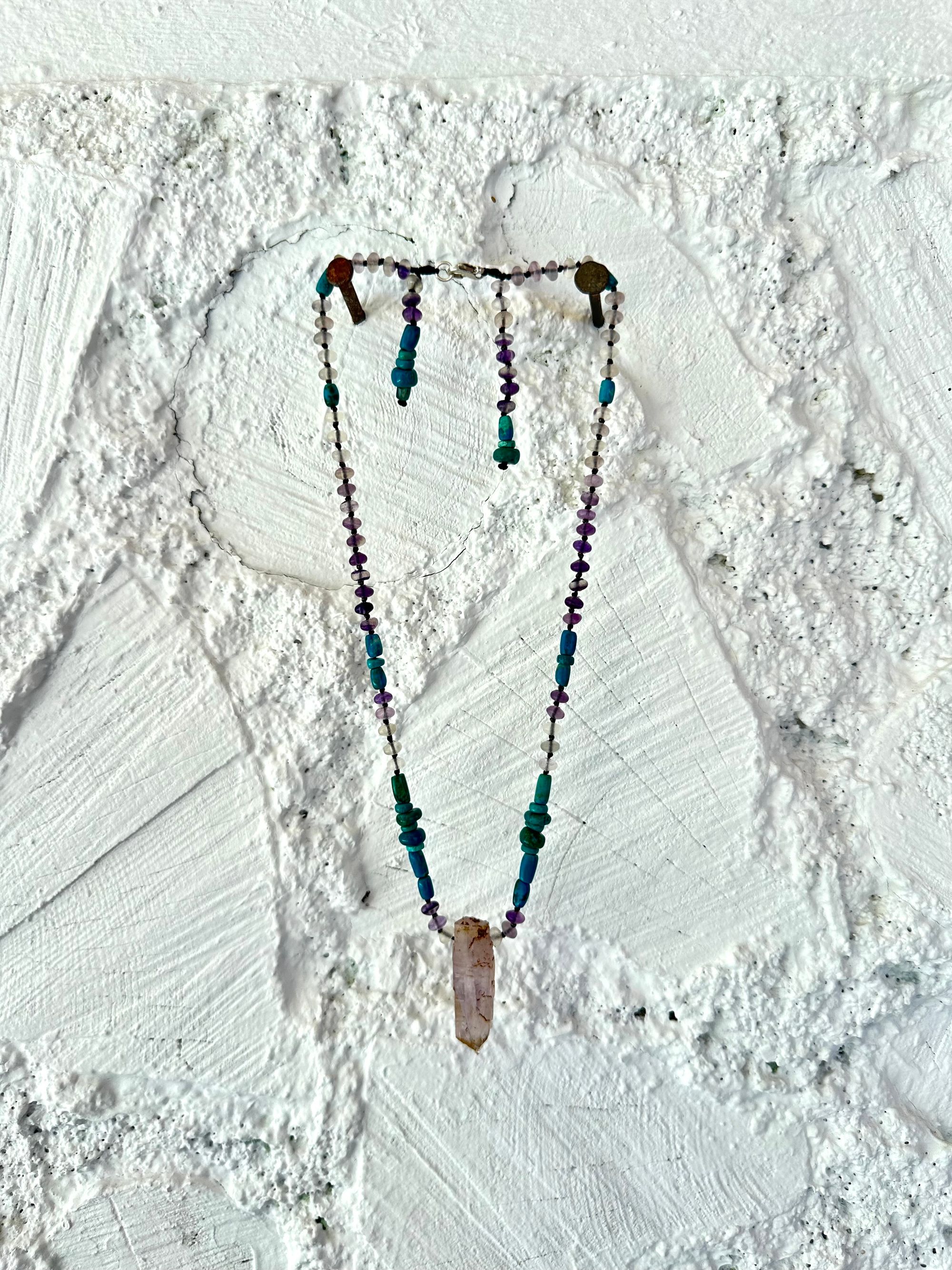 Hanging on a white cordwood wall is a short necklace of soft, purple, amethyst, crystal rich blue green chrysocolla And purple fluorite