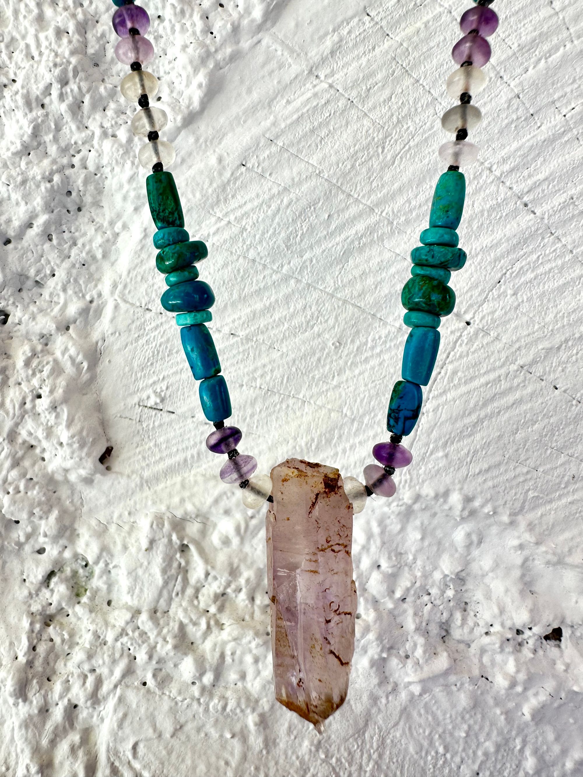 Hanging on a white cordwood wall is a short necklace of soft, purple, amethyst, crystal rich blue green chrysocolla And purple fluorite