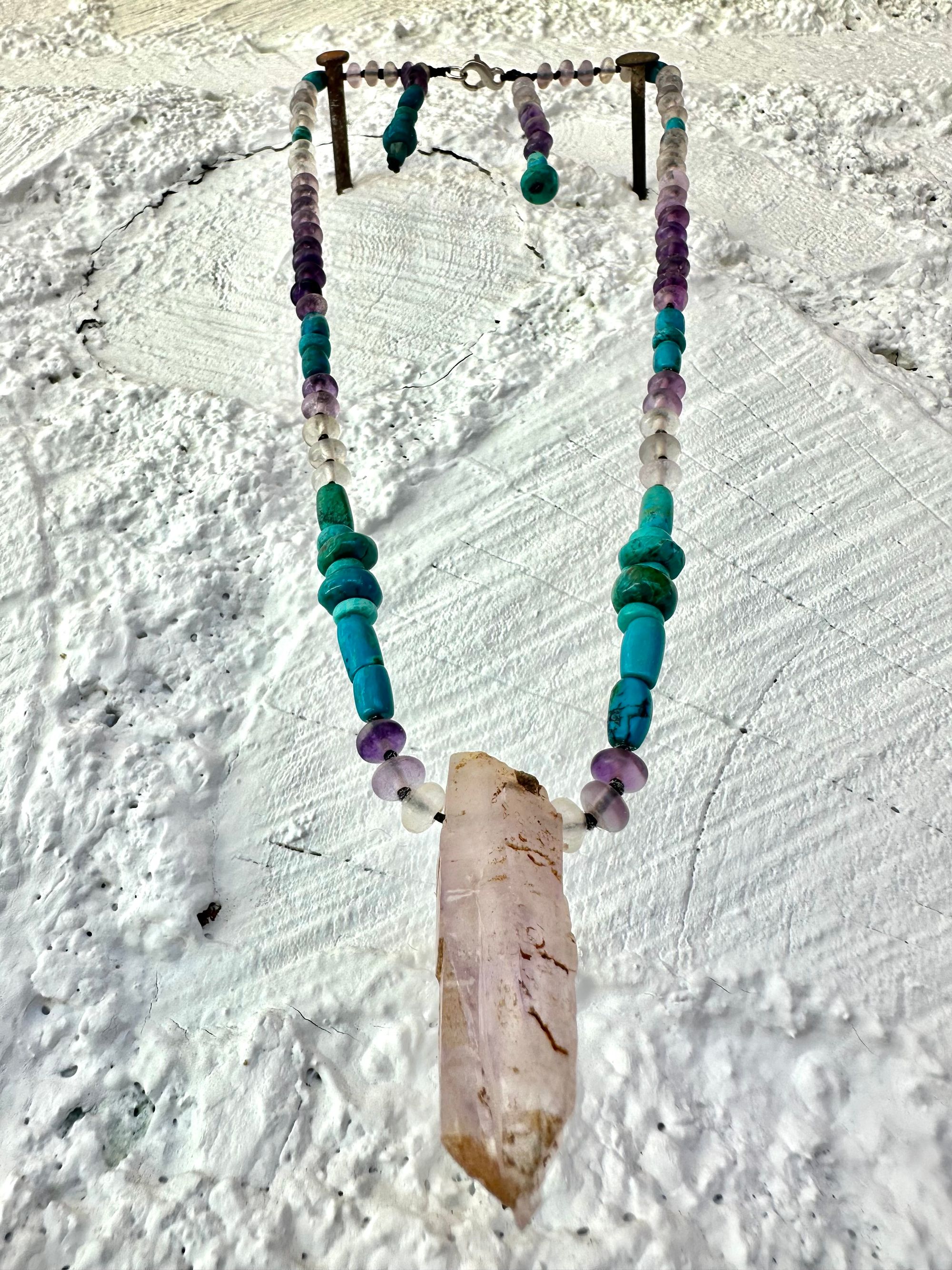 Hanging on a white cordwood wall is a short necklace of soft, purple, amethyst, crystal rich blue green chrysocolla And purple fluorite