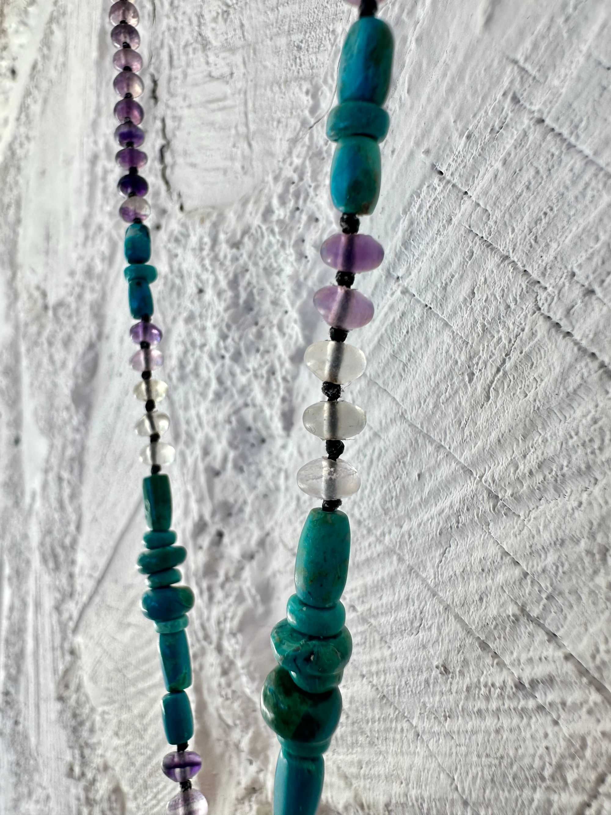 Hanging on a white cordwood wall is a short necklace of soft, purple, amethyst, crystal rich blue green chrysocolla And purple fluorite