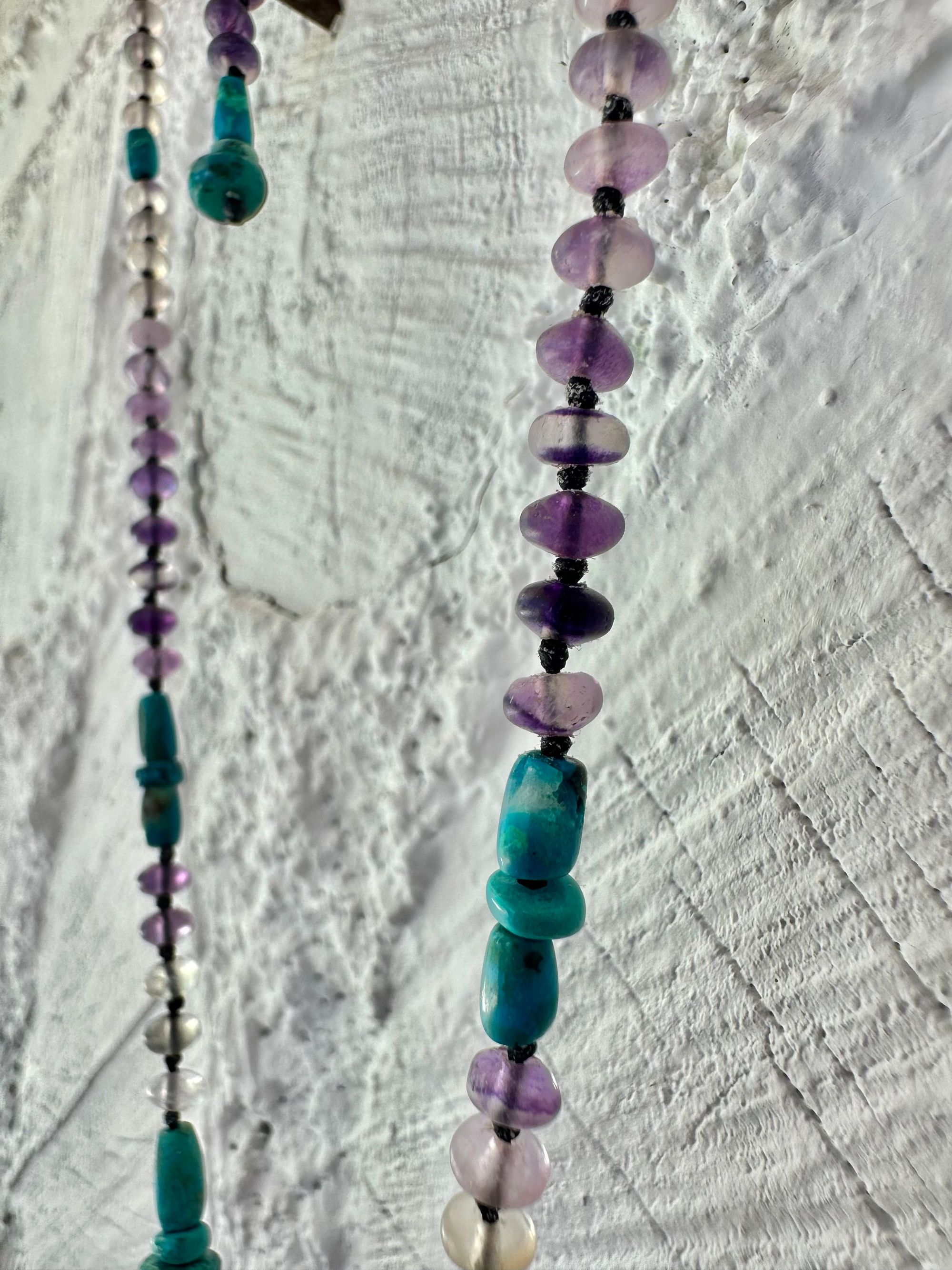 Hanging on a white cordwood wall is a short necklace of soft, purple, amethyst, crystal rich blue green chrysocolla And purple fluorite