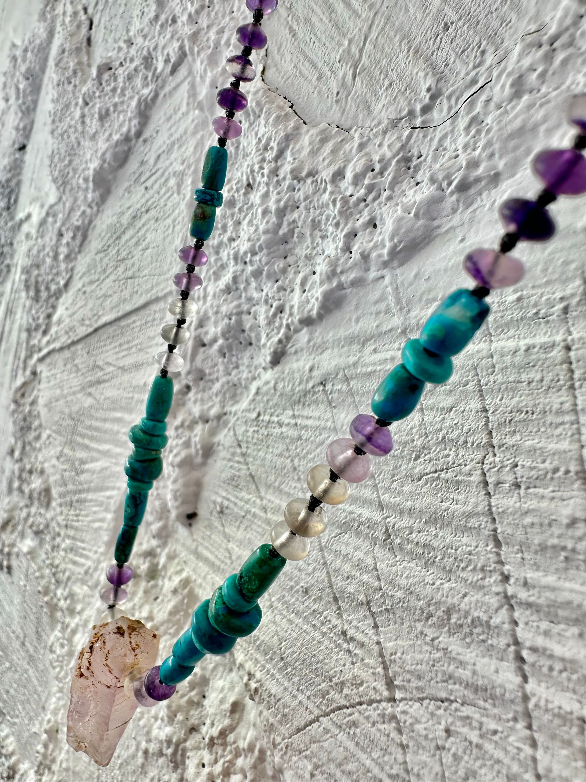 Hanging on a white cordwood wall is a short necklace of soft, purple, amethyst, crystal rich blue green chrysocolla And purple fluorite