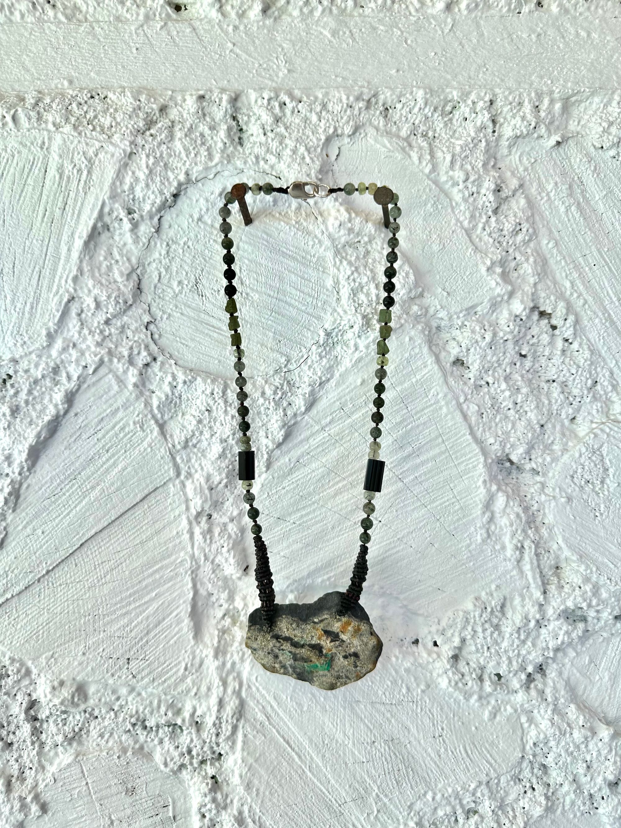 In necklace of raw emerald in its matrix with glittering pyrite is hanging from strands of delicate black work with green beads of rutile, quartz, peridot and black tourmaline