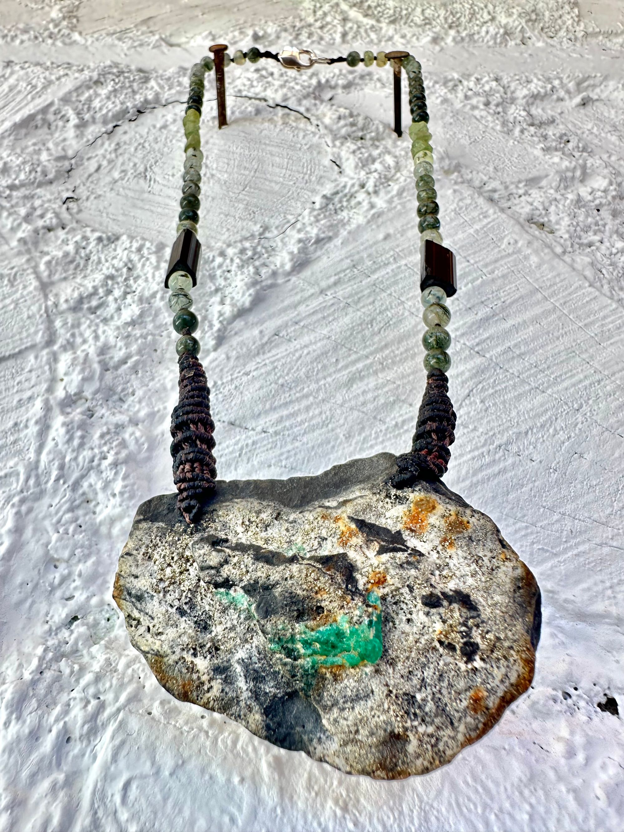 In necklace of raw emerald in its matrix with glittering pyrite is hanging from strands of delicate black work with green beads of rutile, quartz, peridot and black tourmaline