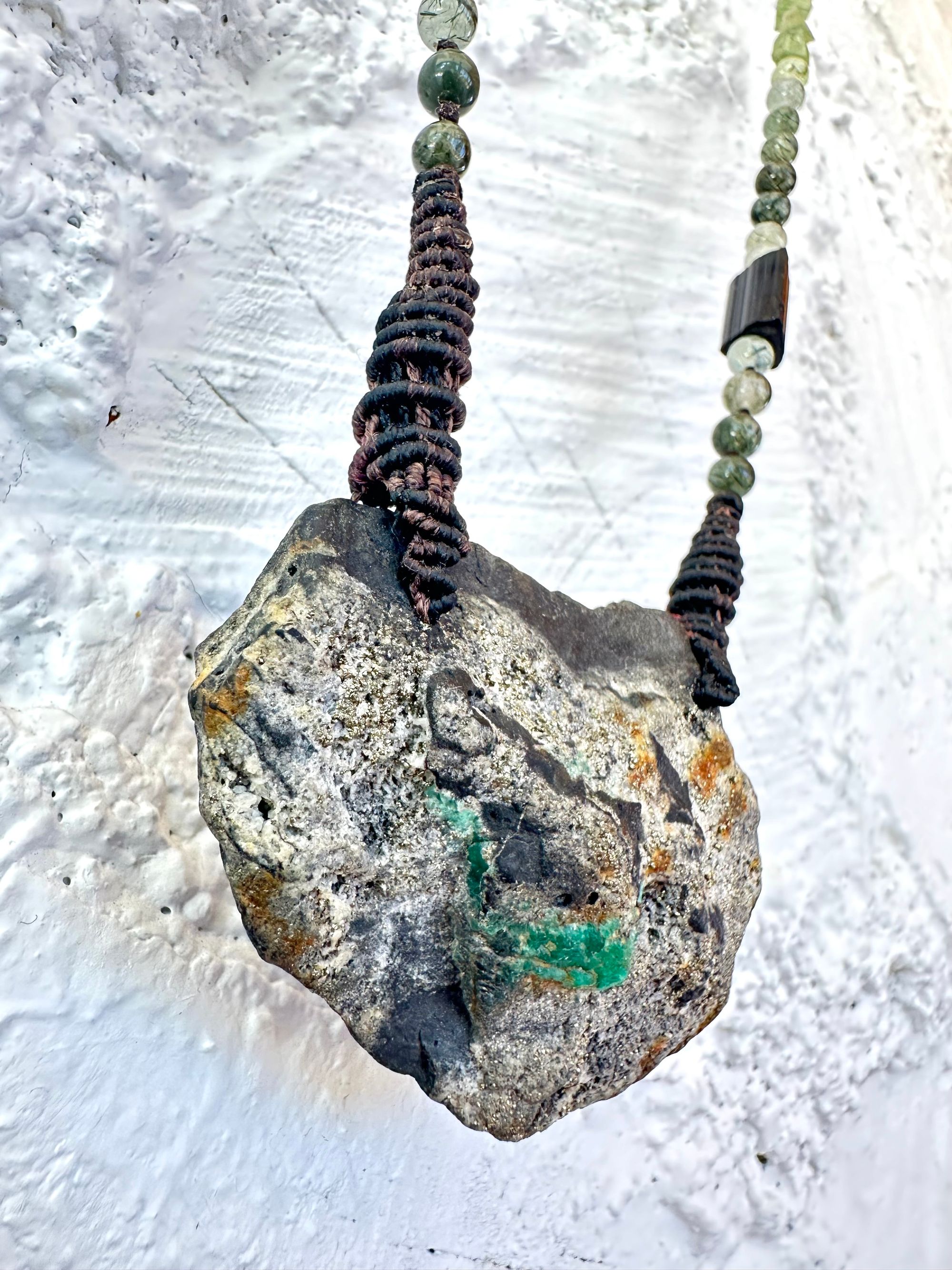 In necklace of raw emerald in its matrix with glittering pyrite is hanging from strands of delicate black work with green beads of rutile, quartz, peridot and black tourmaline