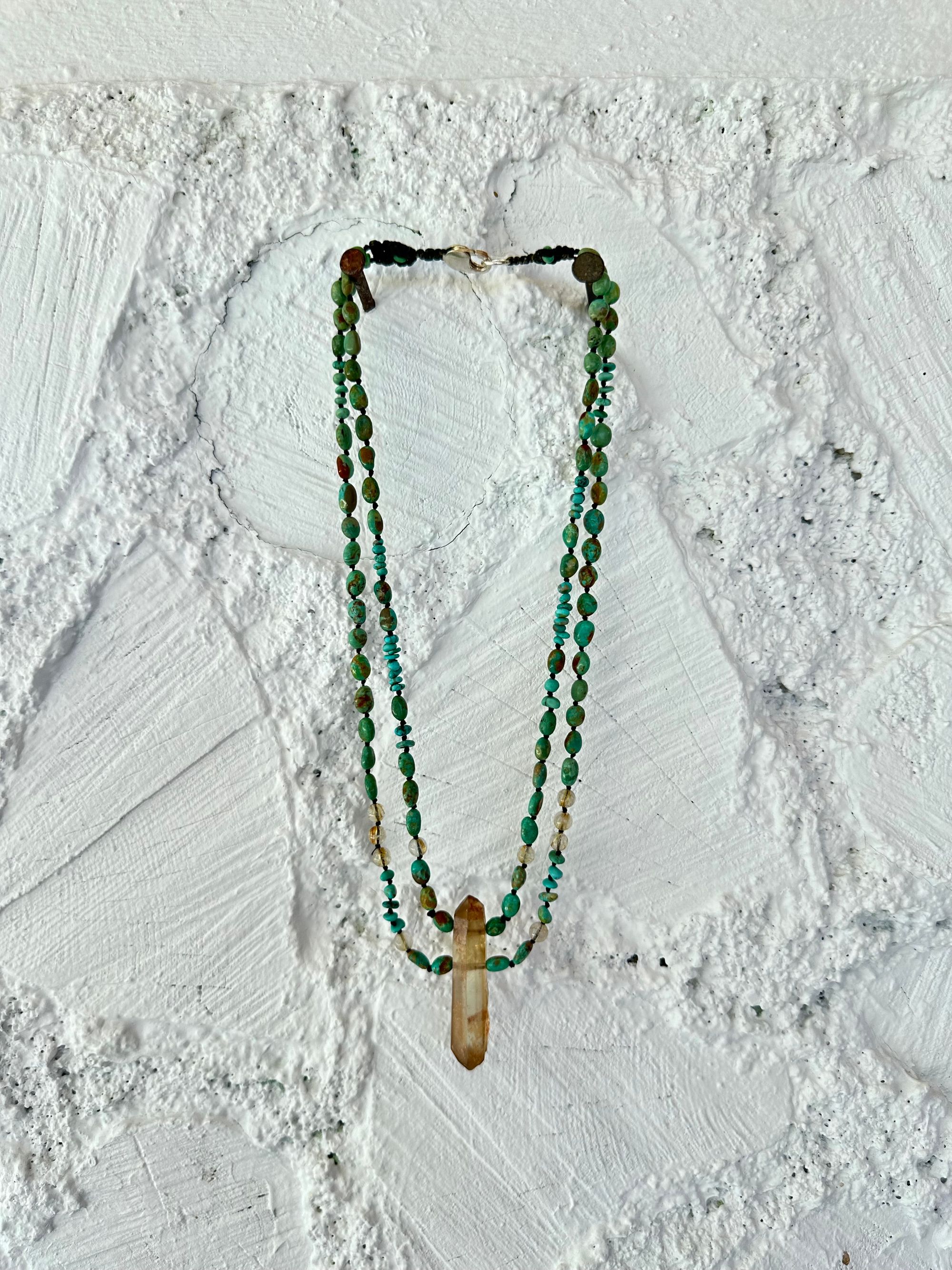 A richly hued Crystal of golden brown citrine Hangs from two strands of deep blue, green, turquoise and gold rutile quartz against a white painted wall of cordwood