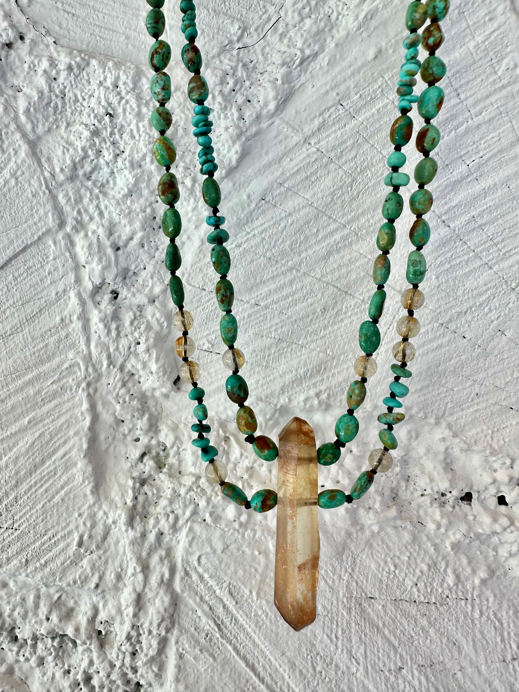 A richly hued Crystal of golden brown citrine Hangs from two strands of deep blue, green, turquoise and gold rutile quartz against a white painted wall of cordwood