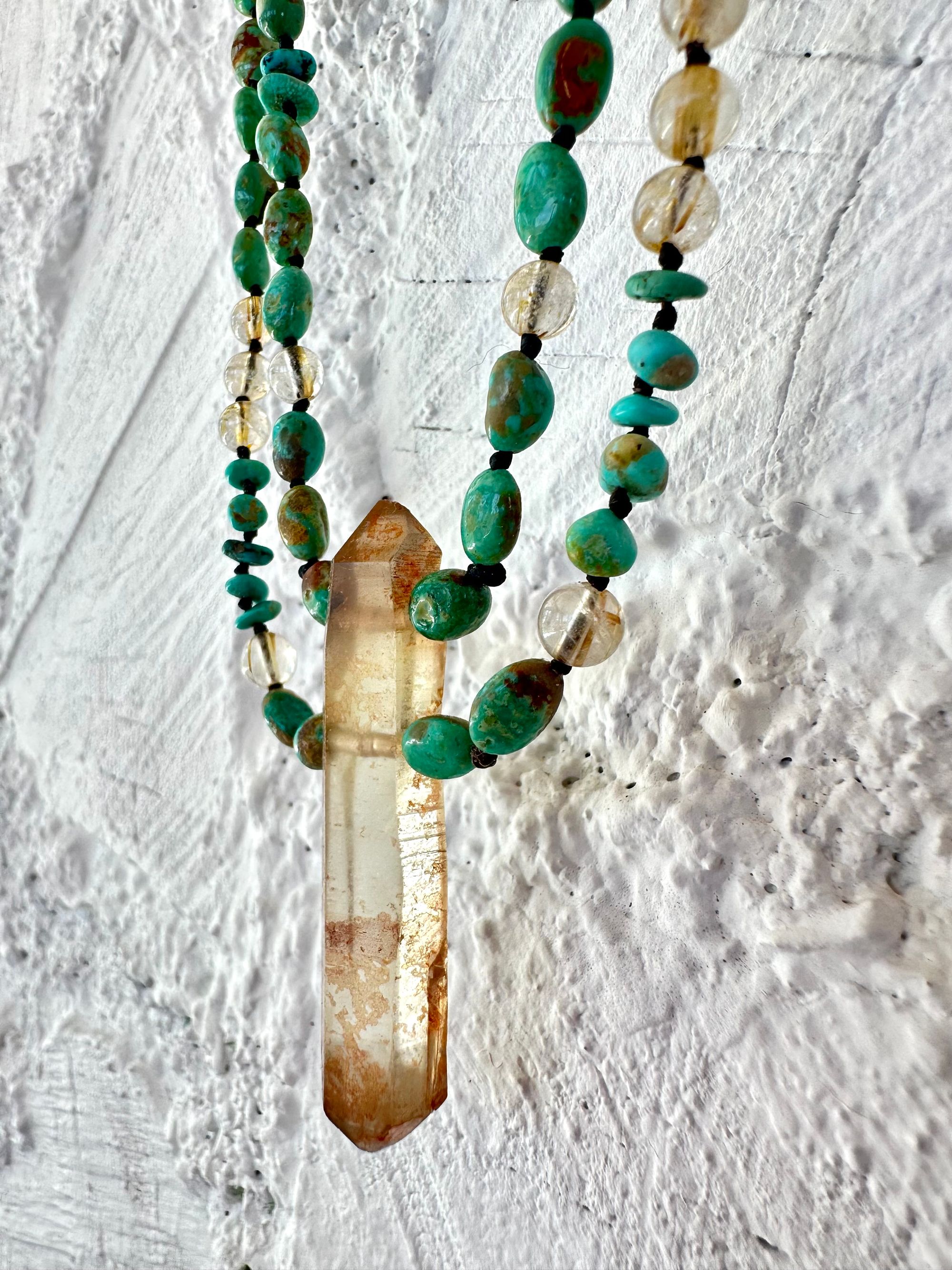 A richly hued Crystal of golden brown citrine Hangs from two strands of deep blue, green, turquoise and gold rutile quartz against a white painted wall of cordwood