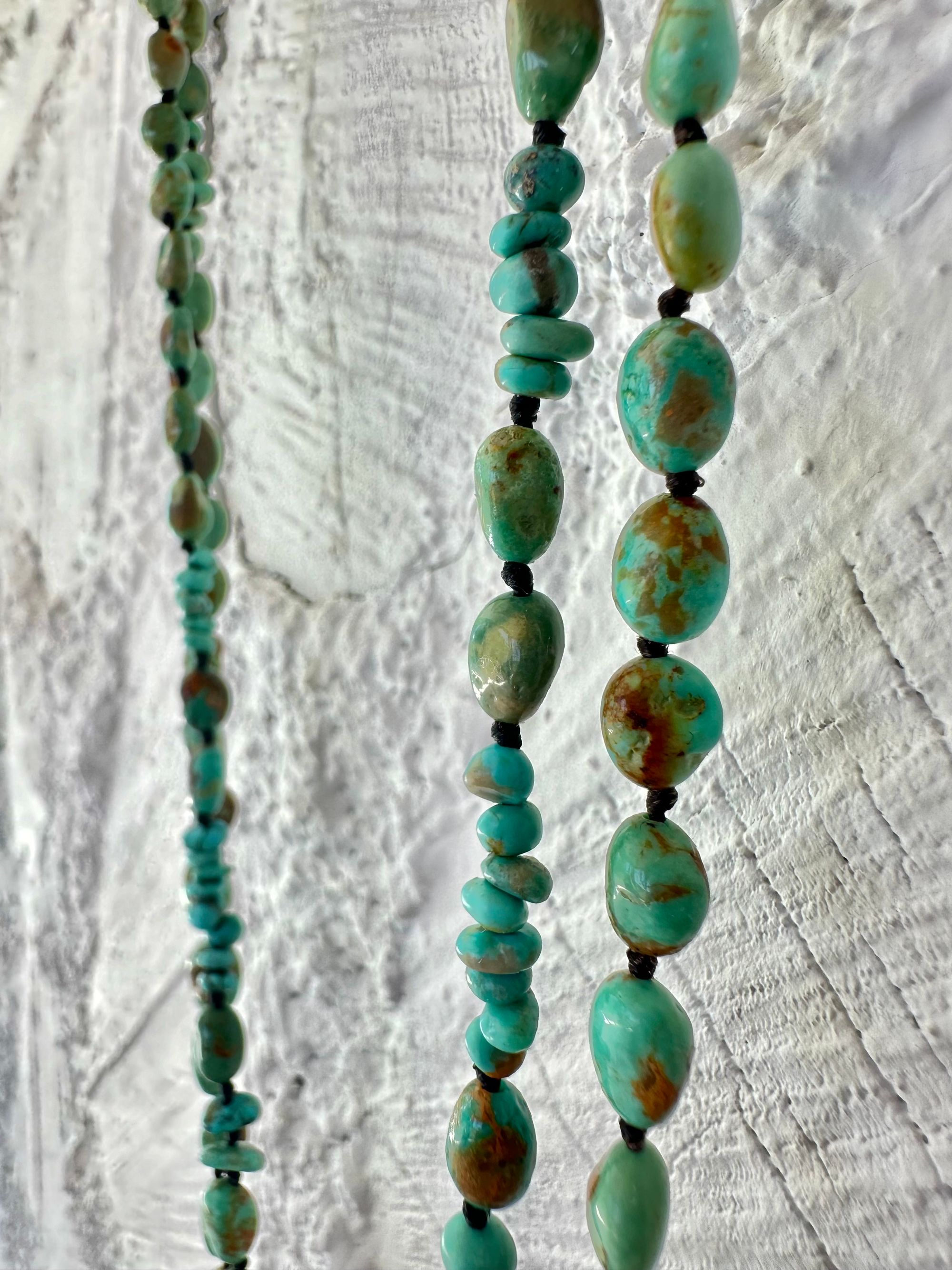 A richly hued Crystal of golden brown citrine Hangs from two strands of deep blue, green, turquoise and gold rutile quartz against a white painted wall of cordwood