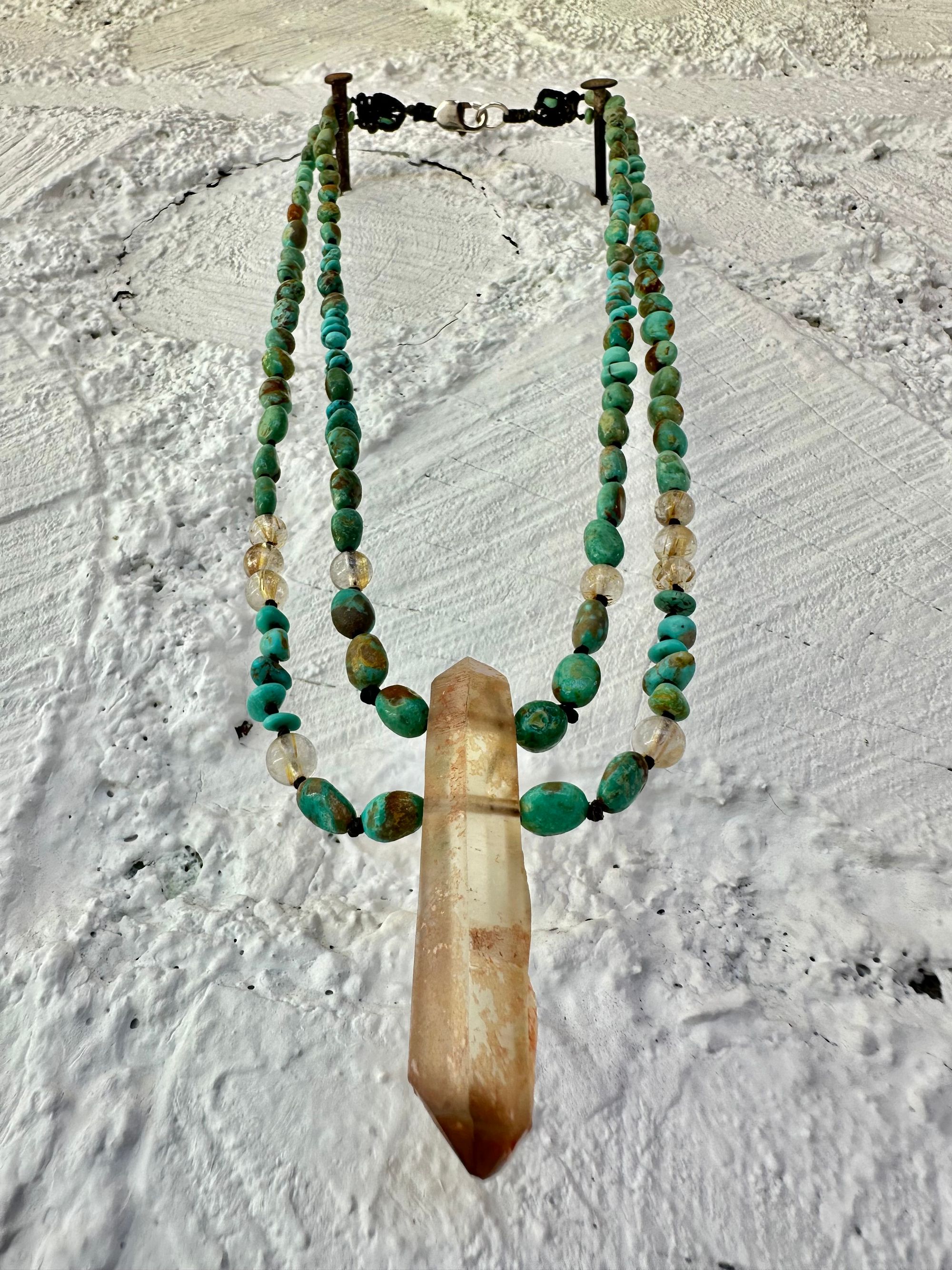 A richly hued Crystal of golden brown citrine Hangs from two strands of deep blue, green, turquoise and gold rutile quartz against a white painted wall of cordwood