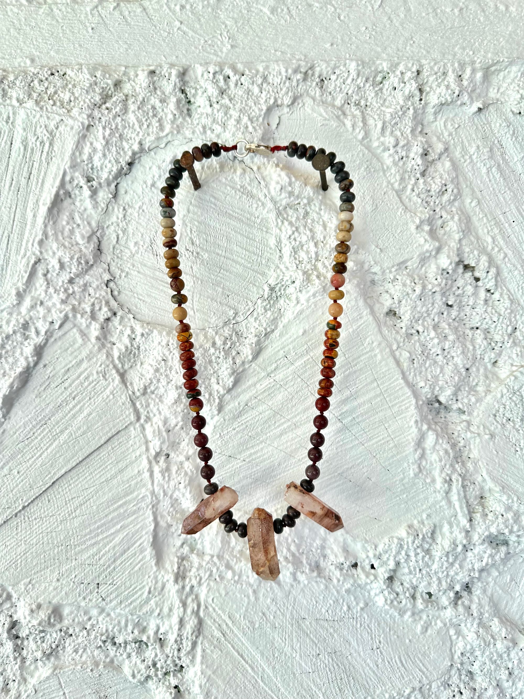 A necklace With three red iron quartz points and a rainbow of oval picture, Jasper beads hangs on a white cordwood wall. The Jasper flows in gradations from deep red black to fiery, red, orange and green.