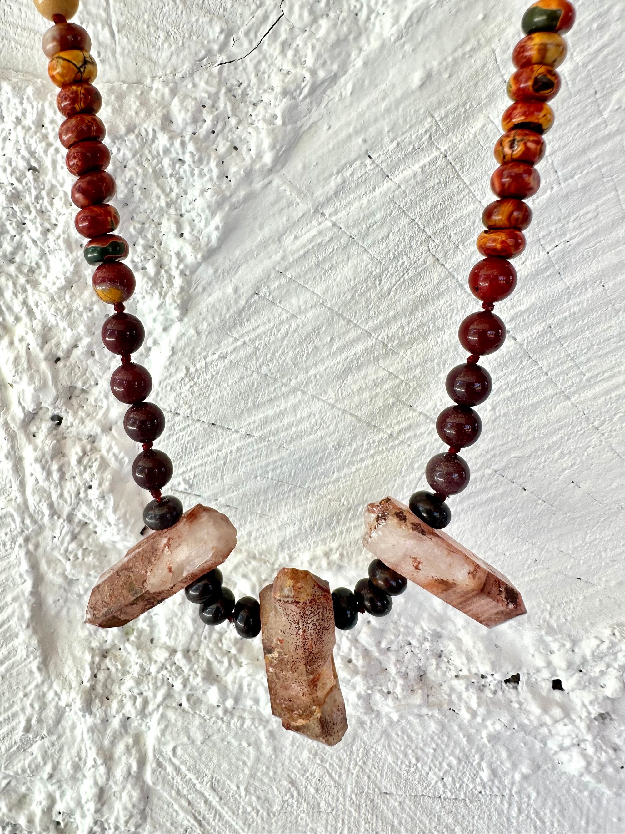 A necklace With three red iron quartz points and a rainbow of oval picture, Jasper beads hangs on a white cordwood wall. The Jasper flows in gradations from deep red black to fiery, red, orange and green.