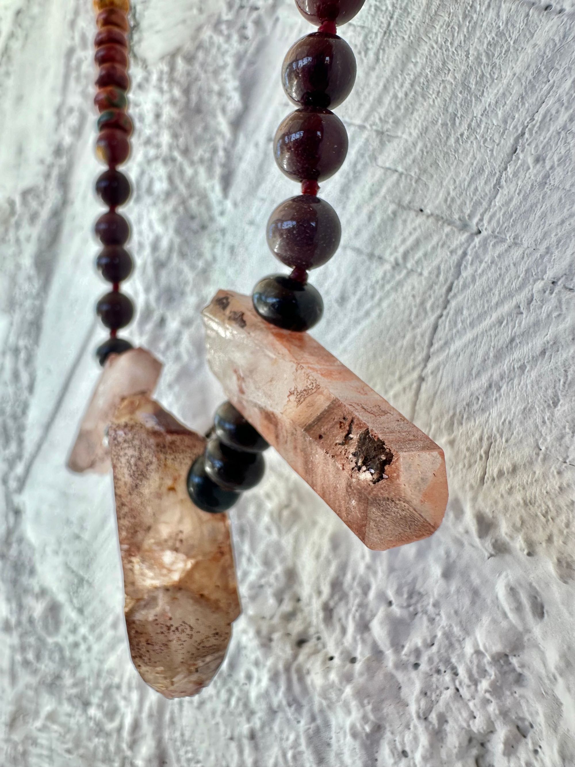 A necklace With three red iron quartz points and a rainbow of oval picture, Jasper beads hangs on a white cordwood wall. The Jasper flows in gradations from deep red black to fiery, red, orange and green.