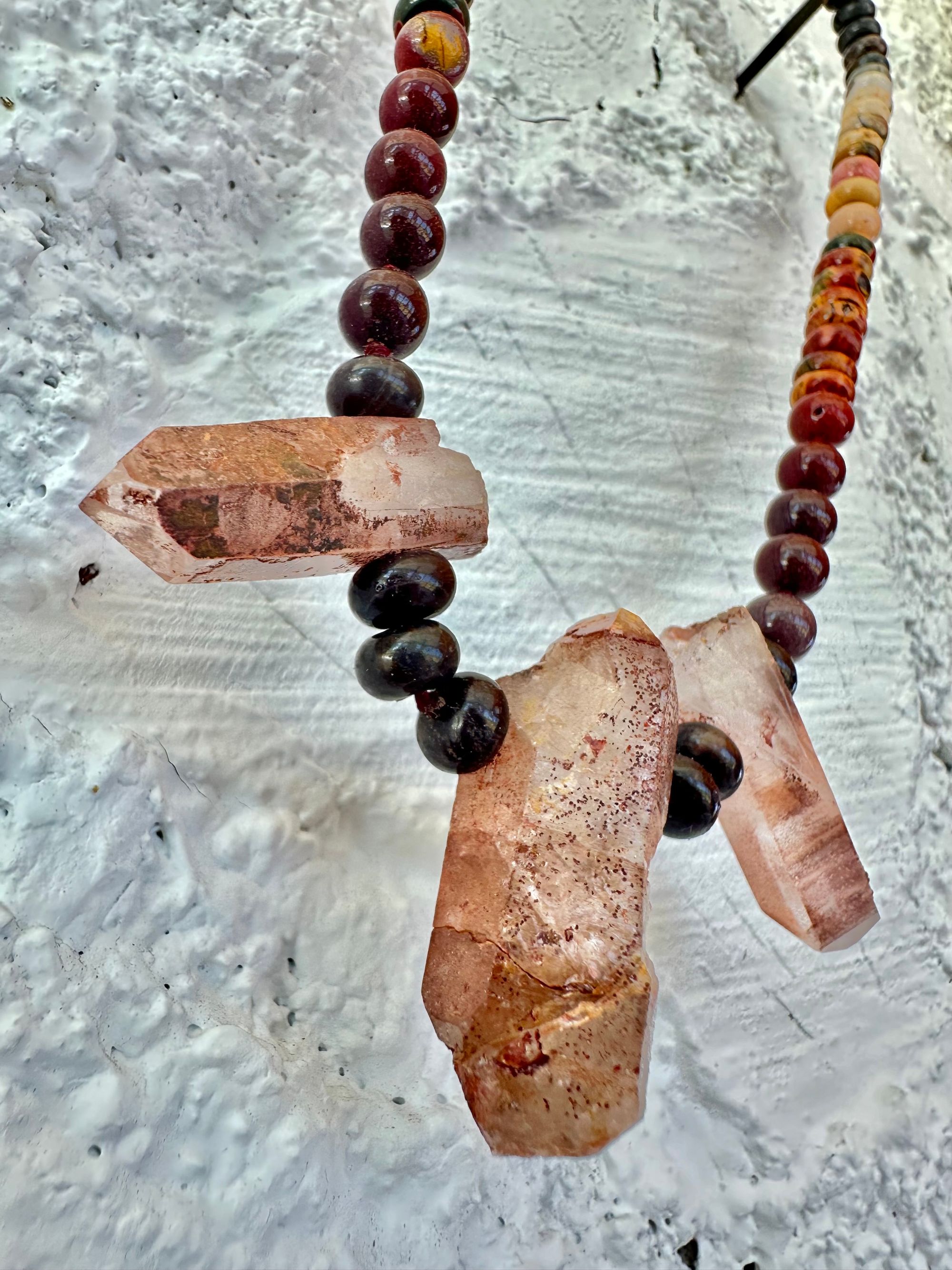 A necklace With three red iron quartz points and a rainbow of oval picture, Jasper beads hangs on a white cordwood wall. The Jasper flows in gradations from deep red black to fiery, red, orange and green.