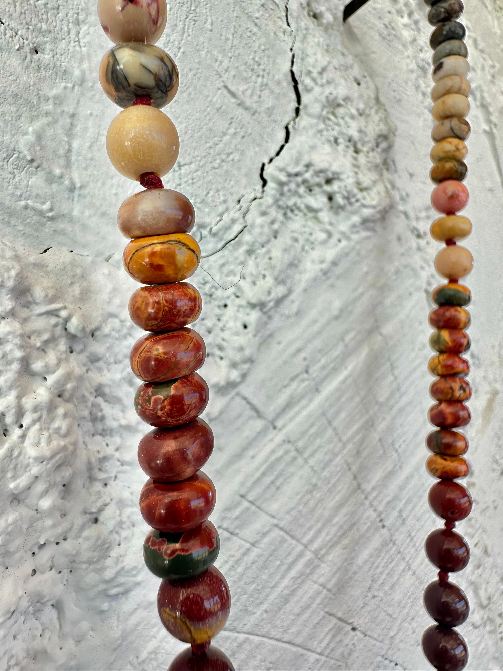 A necklace With three red iron quartz points and a rainbow of oval picture, Jasper beads hangs on a white cordwood wall. The Jasper flows in gradations from deep red black to fiery, red, orange and green.