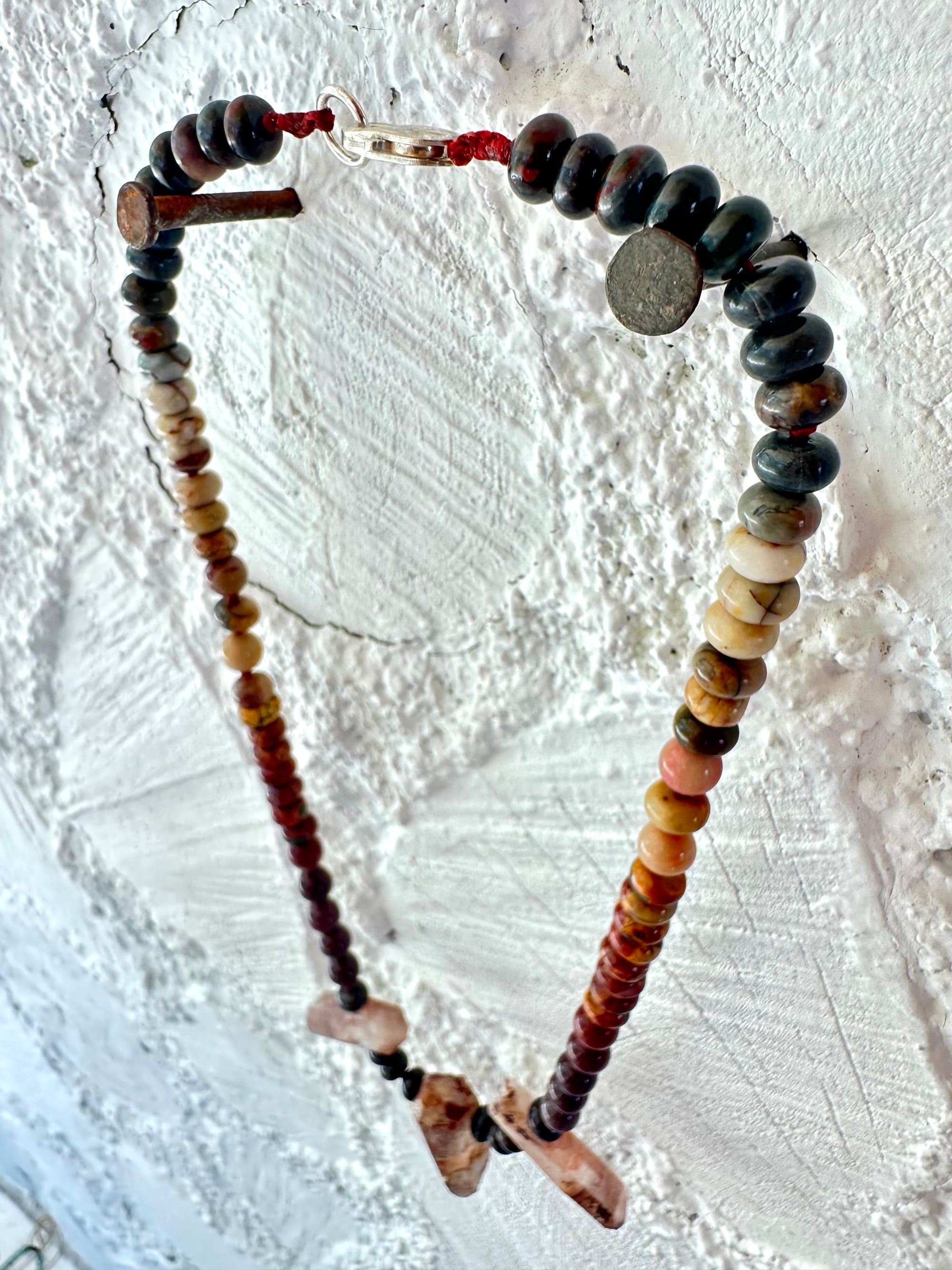 A necklace With three red iron quartz points and a rainbow of oval picture, Jasper beads hangs on a white cordwood wall. The Jasper flows in gradations from deep red black to fiery, red, orange and green.