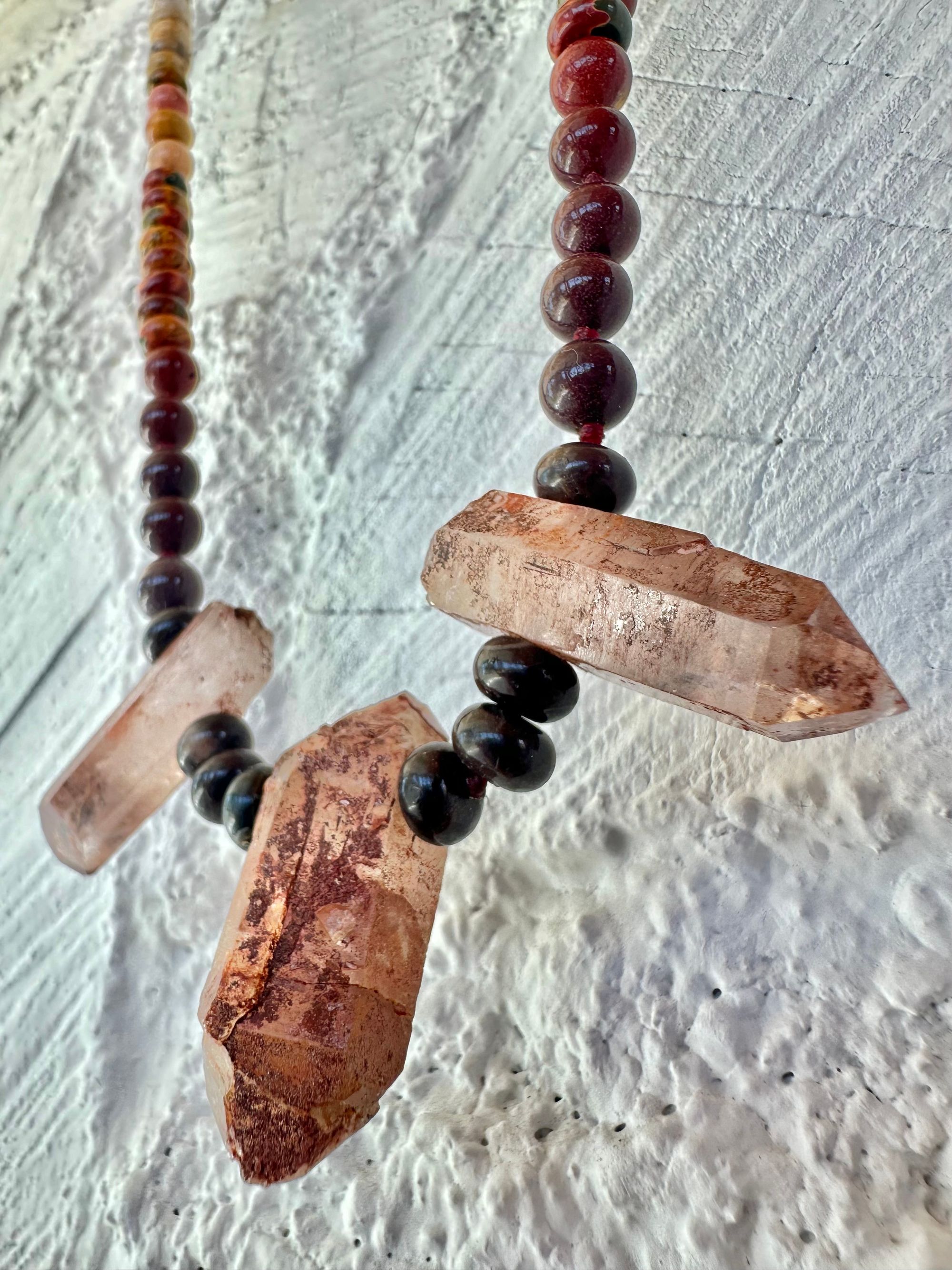 A necklace With three red iron quartz points and a rainbow of oval picture, Jasper beads hangs on a white cordwood wall. The Jasper flows in gradations from deep red black to fiery, red, orange and green.