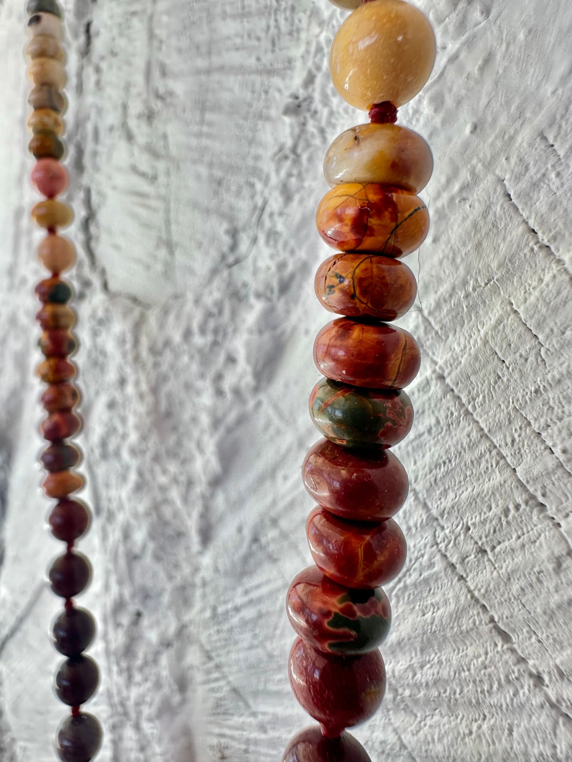 A necklace With three red iron quartz points and a rainbow of oval picture, Jasper beads hangs on a white cordwood wall. The Jasper flows in gradations from deep red black to fiery, red, orange and green.