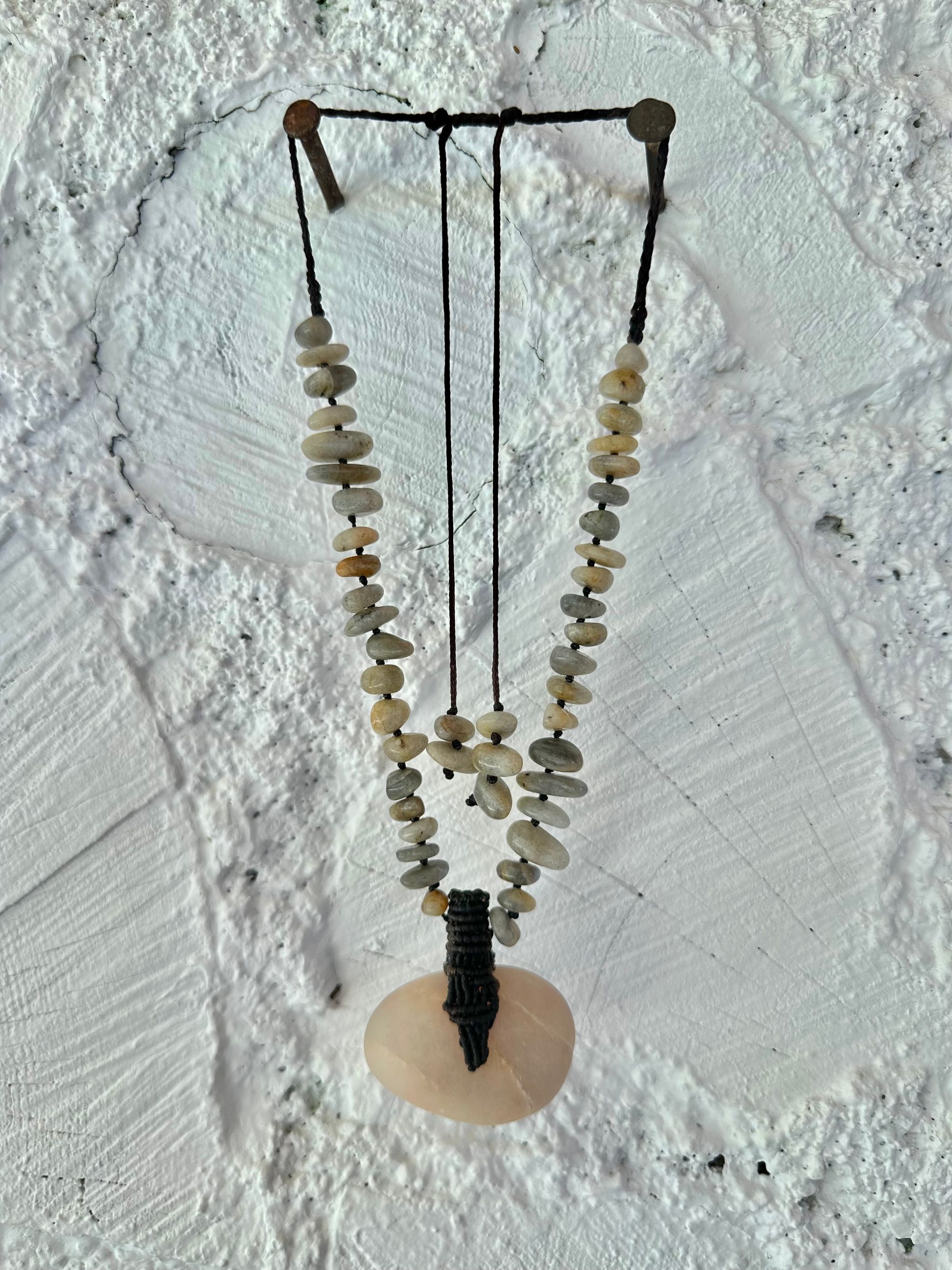 A detail of a necklace constructed from a White glowing ocean tumbled stone, From it was constructed a dark purple, knot work Basket, and from this Are irregularly shaped Beads of labradorite The necklace hangs against a wall of white cordwood