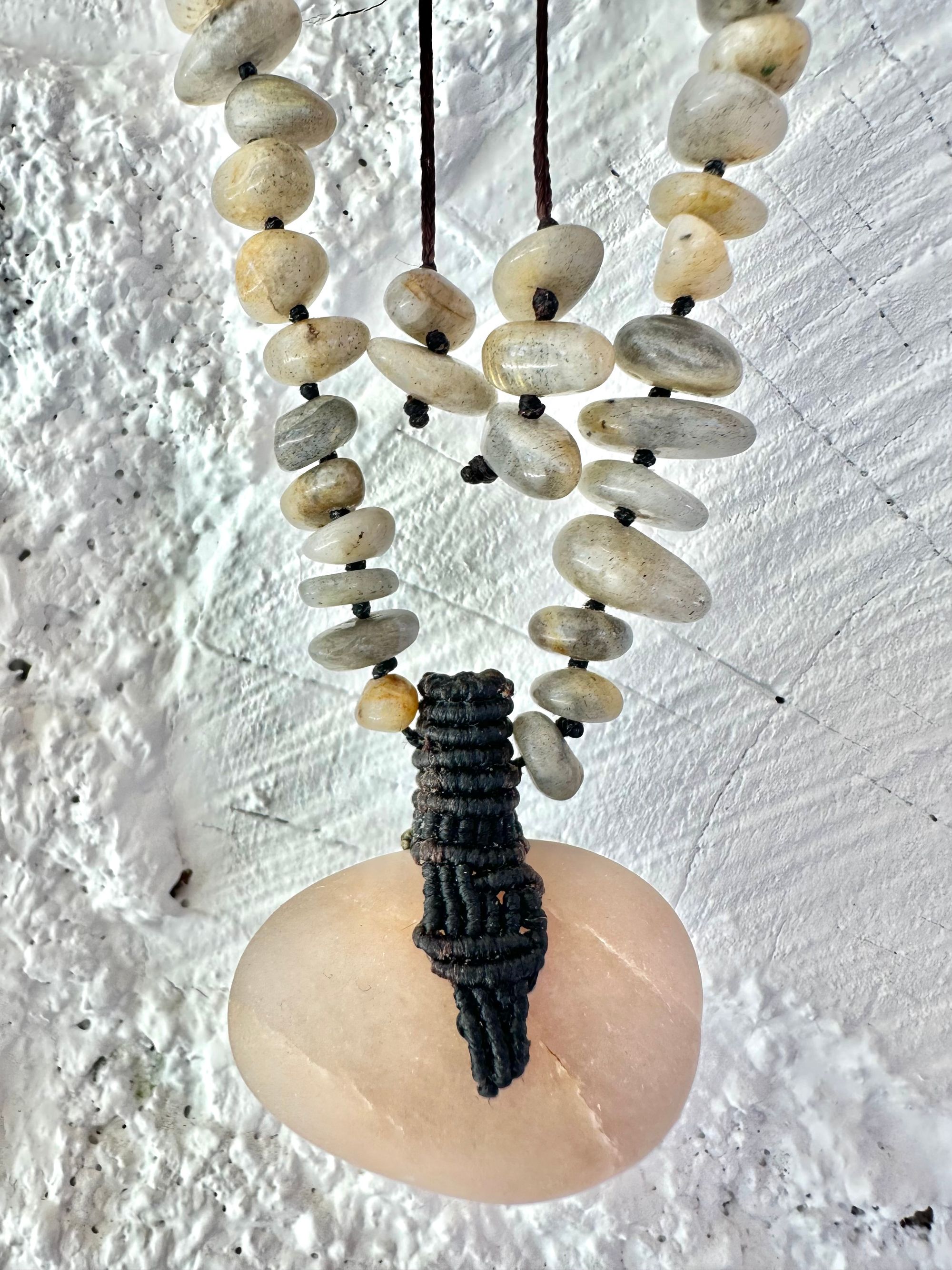 A detail of a necklace constructed from a White glowing ocean tumbled stone, From it was constructed a dark purple, knot work Basket, and from this Are irregularly shaped Beads of labradorite The necklace hangs against a wall of white cordwood