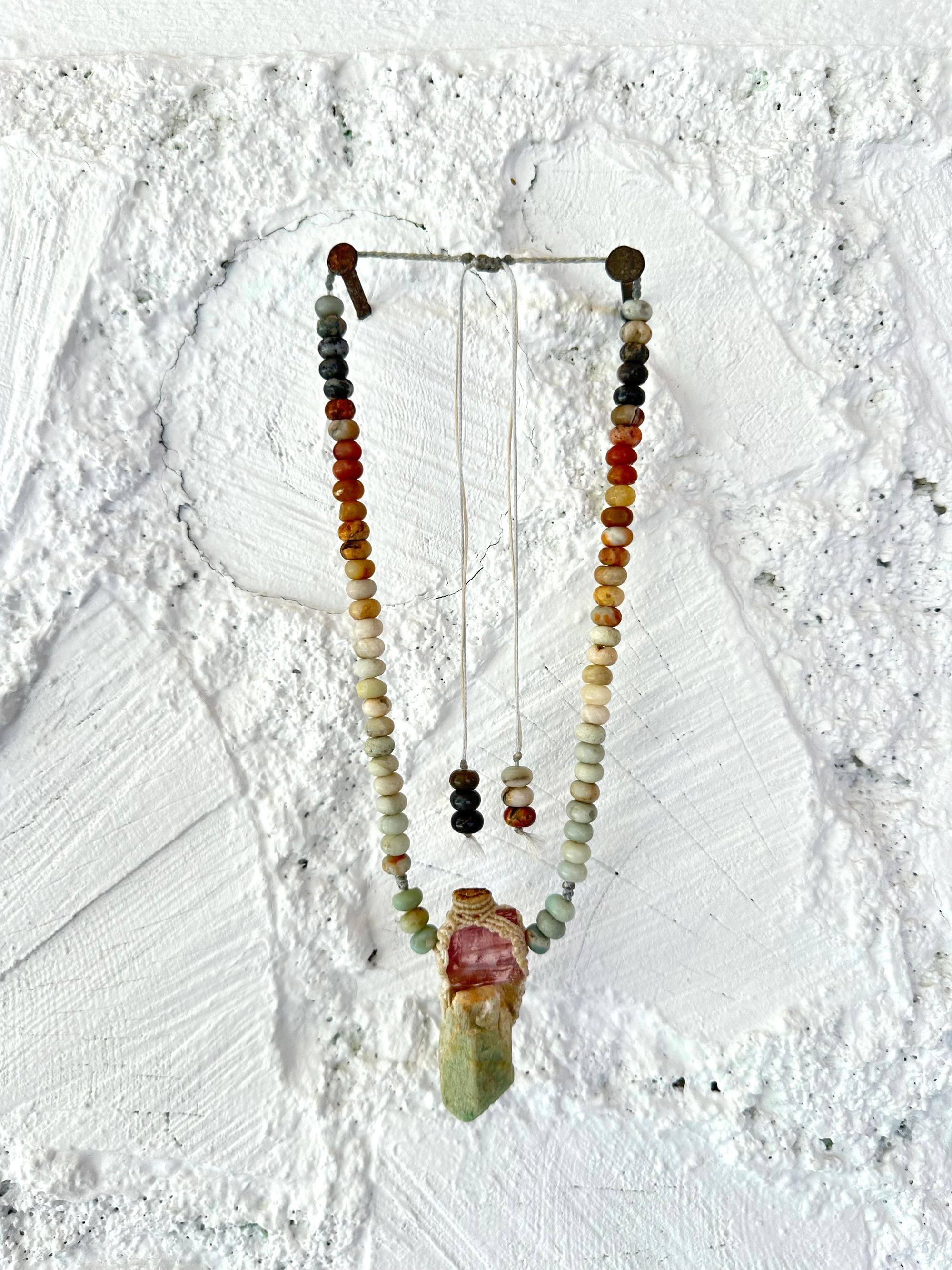 Detail of A luminous, pink tourmaline, soft turquoise amazonite stone And knotwork Basket necklace With beads of grated blue to white to brown amazonite Against a white painted cordwood wall