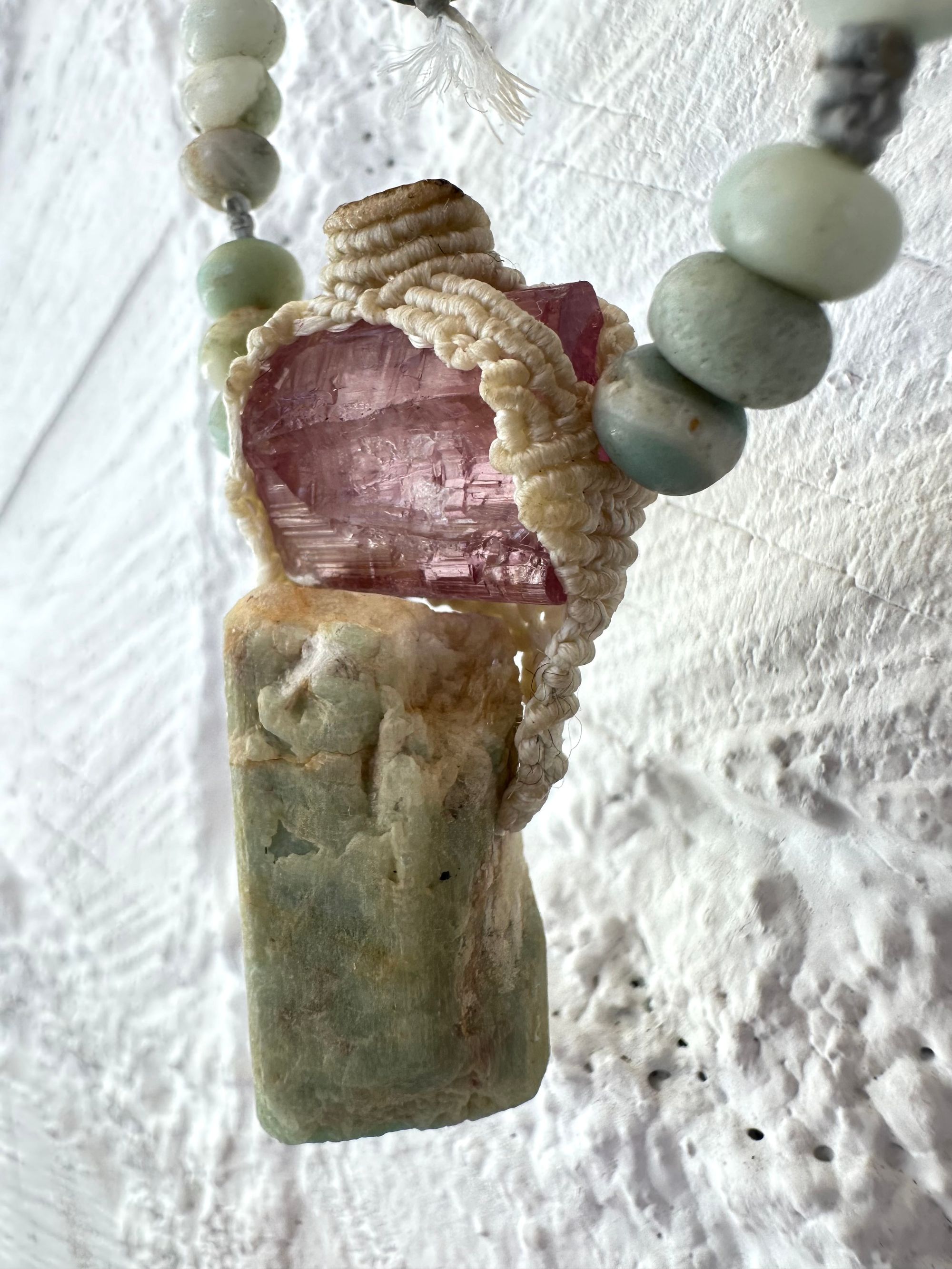 Detail of A luminous, pink tourmaline, soft turquoise amazonite stone And knotwork Basket necklace With beads of grated blue to white to brown amazonite Against a white painted cordwood wall