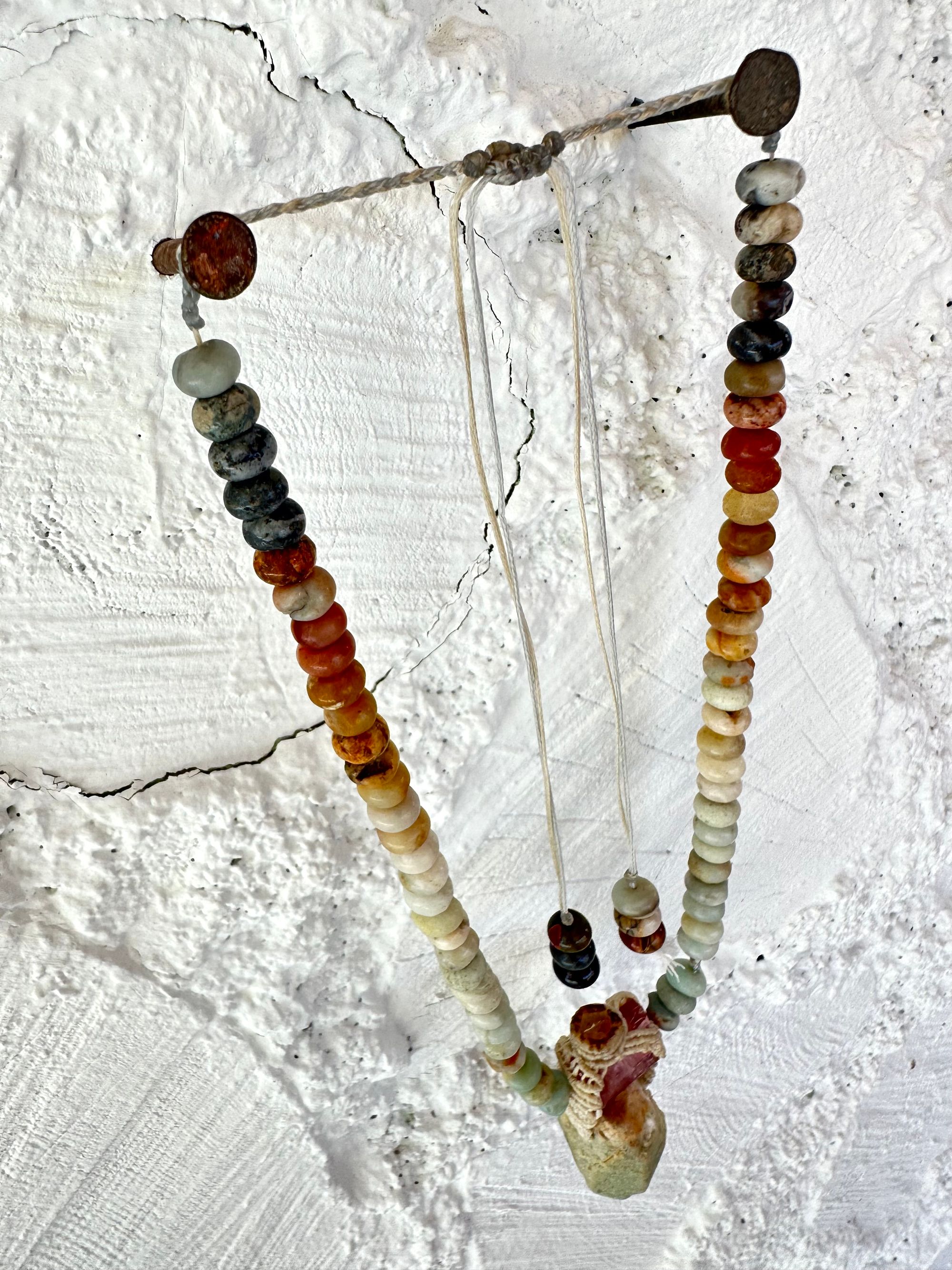 Detail of A luminous, pink tourmaline, soft turquoise amazonite stone And knotwork Basket necklace With beads of grated blue to white to brown amazonite Against a white painted cordwood wall