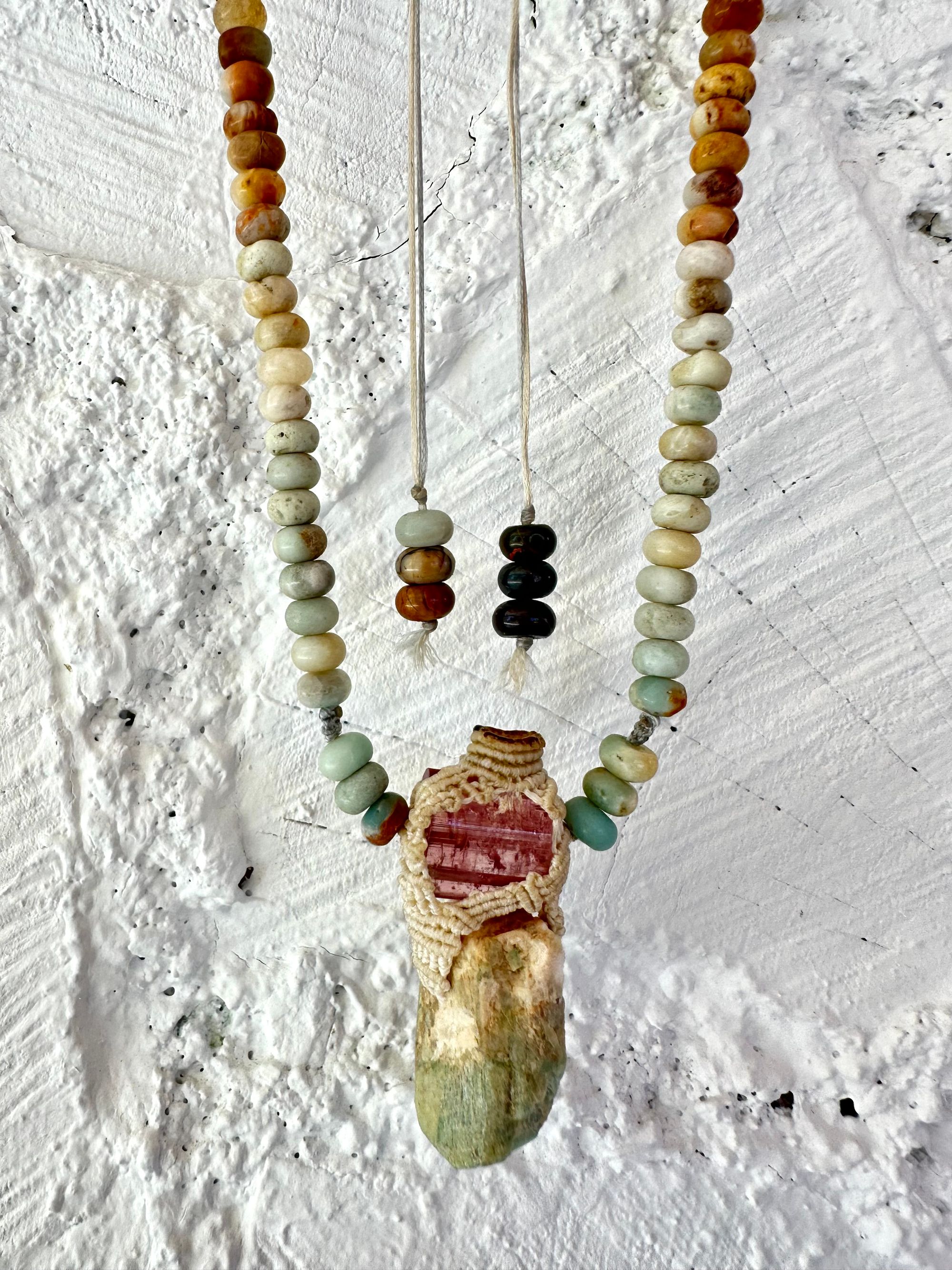 Detail of A luminous, pink tourmaline, soft turquoise amazonite stone And knotwork Basket necklace With beads of grated blue to white to brown amazonite Against a white painted cordwood wall