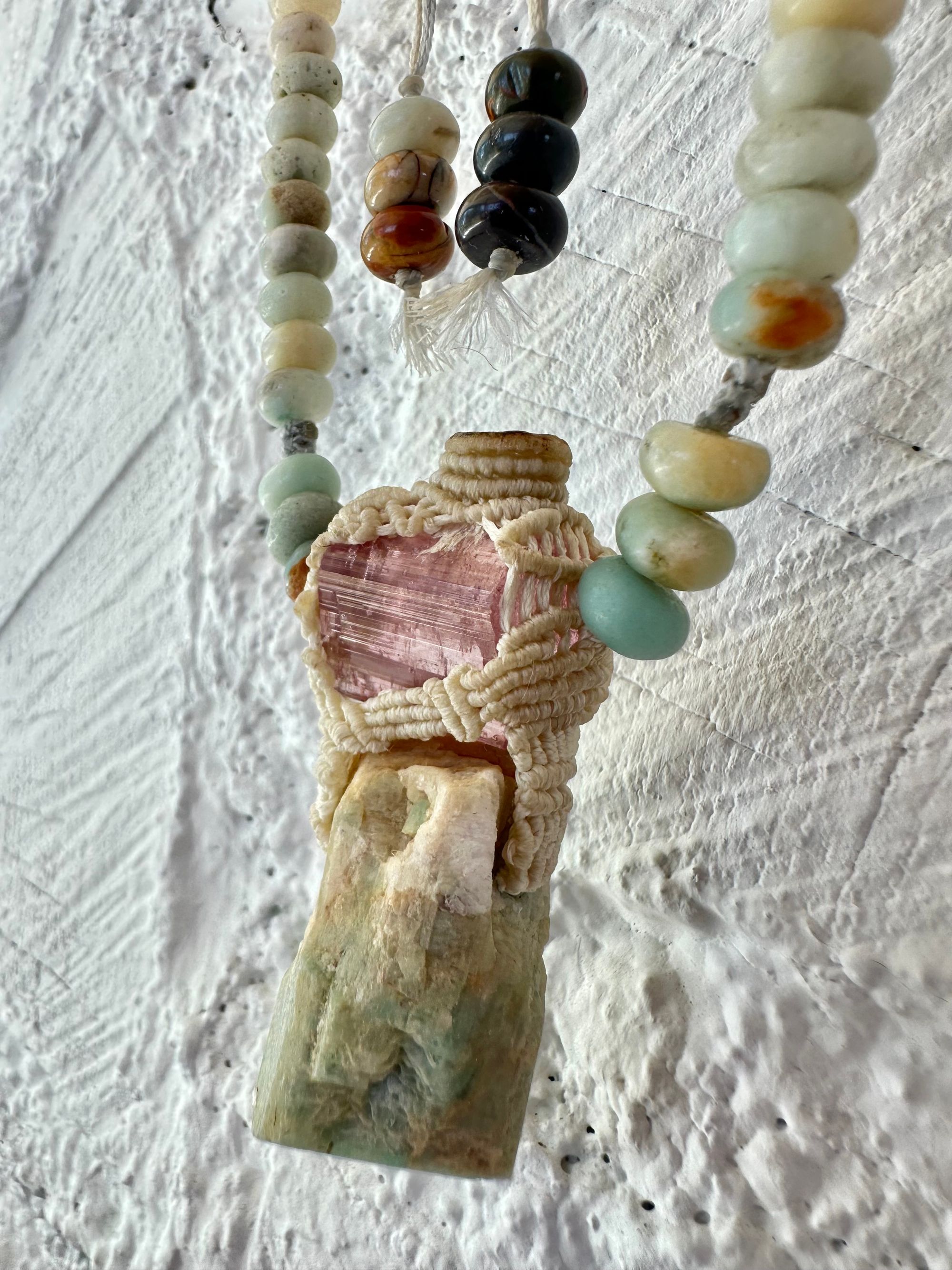 Detail of A luminous, pink tourmaline, soft turquoise amazonite stone And knotwork Basket necklace With beads of grated blue to white to brown amazonite Against a white painted cordwood wall