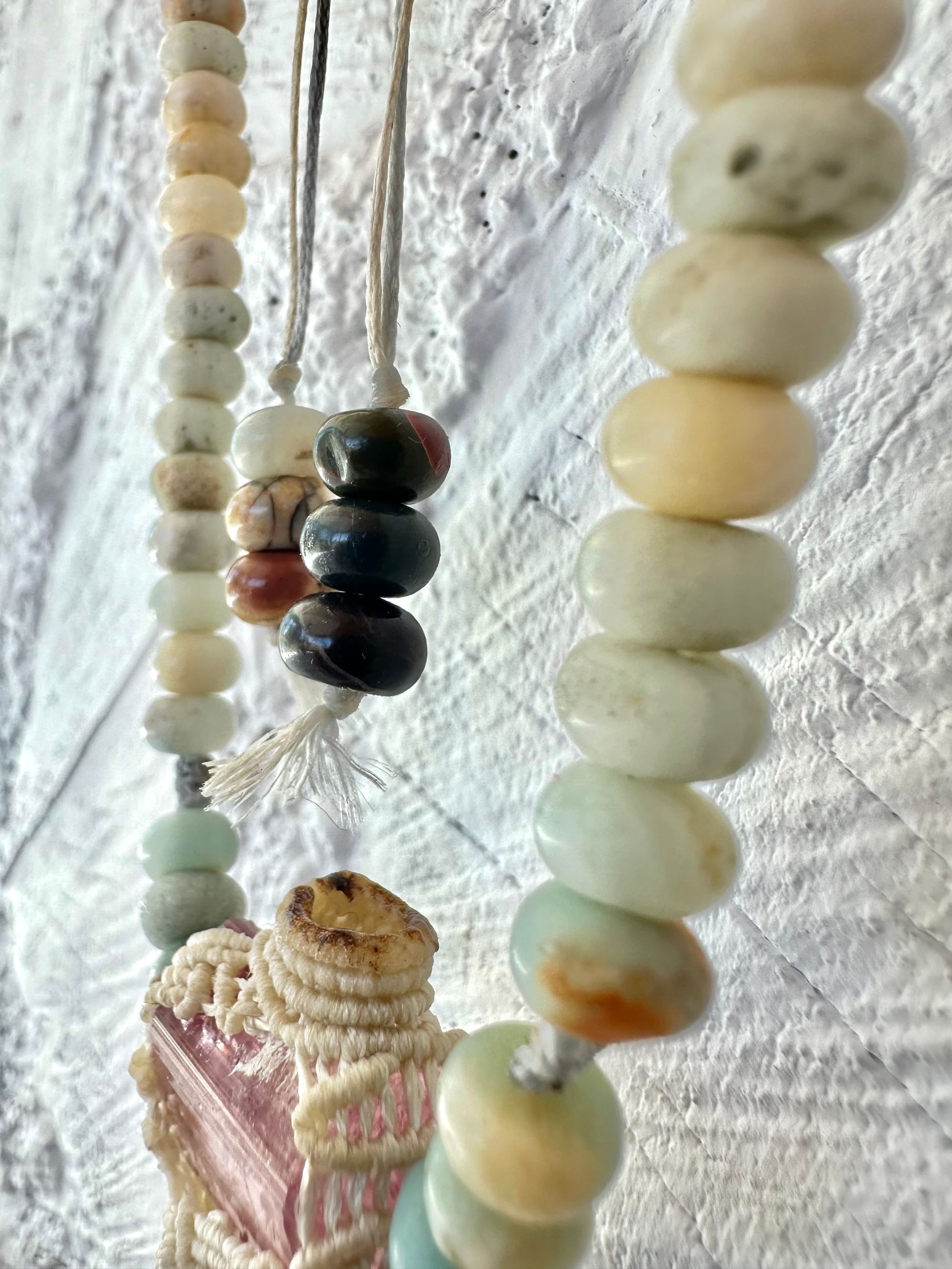 Detail of A luminous, pink tourmaline, soft turquoise amazonite stone And knotwork Basket necklace With beads of grated blue to white to brown amazonite Against a white painted cordwood wall