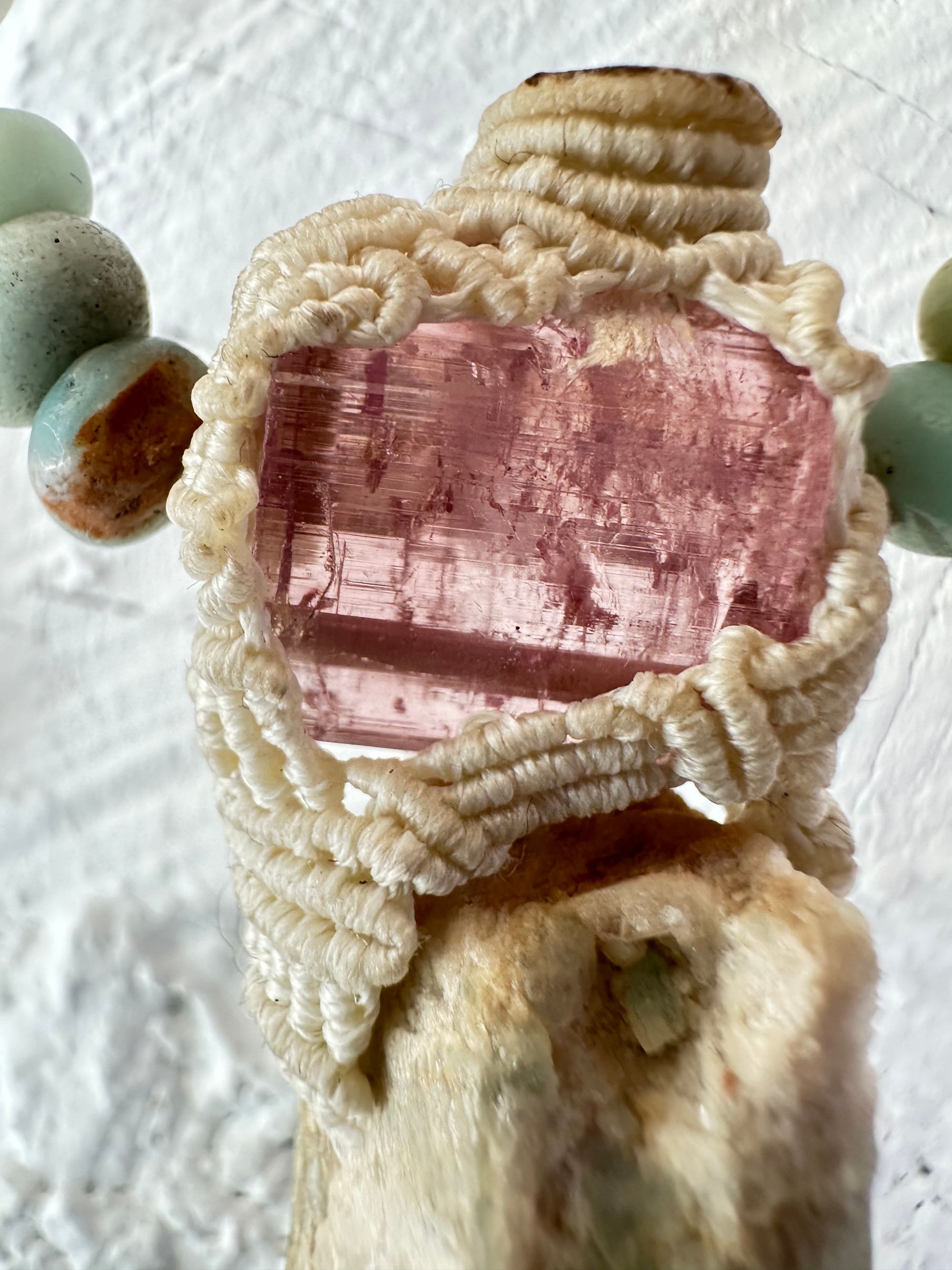 Detail of A luminous, pink tourmaline, soft turquoise amazonite stone And knotwork Basket necklace With beads of grated blue to white to brown amazonite Against a white painted cordwood wall