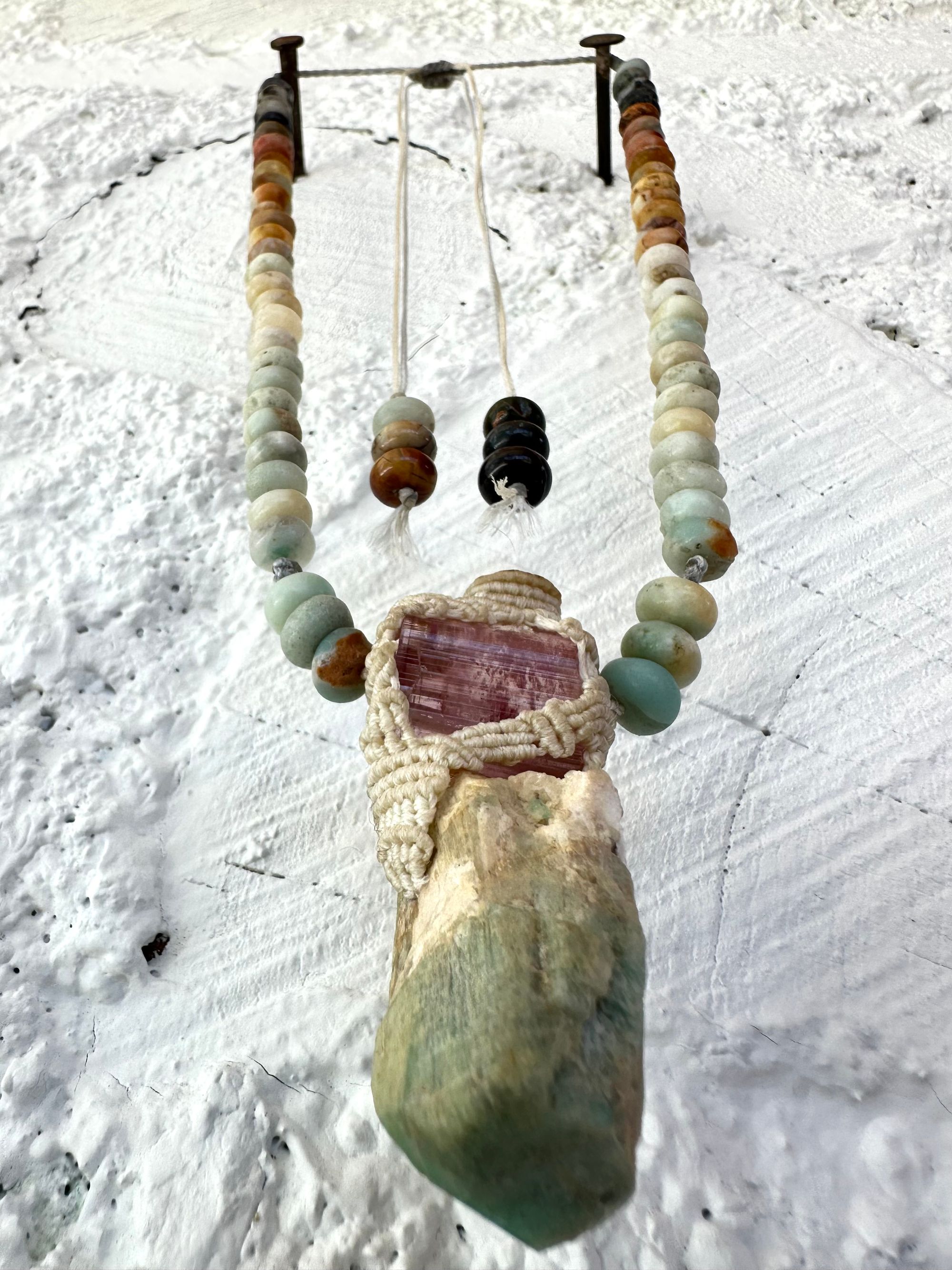 Detail of A luminous, pink tourmaline, soft turquoise amazonite stone And knotwork Basket necklace With beads of grated blue to white to brown amazonite Against a white painted cordwood wall