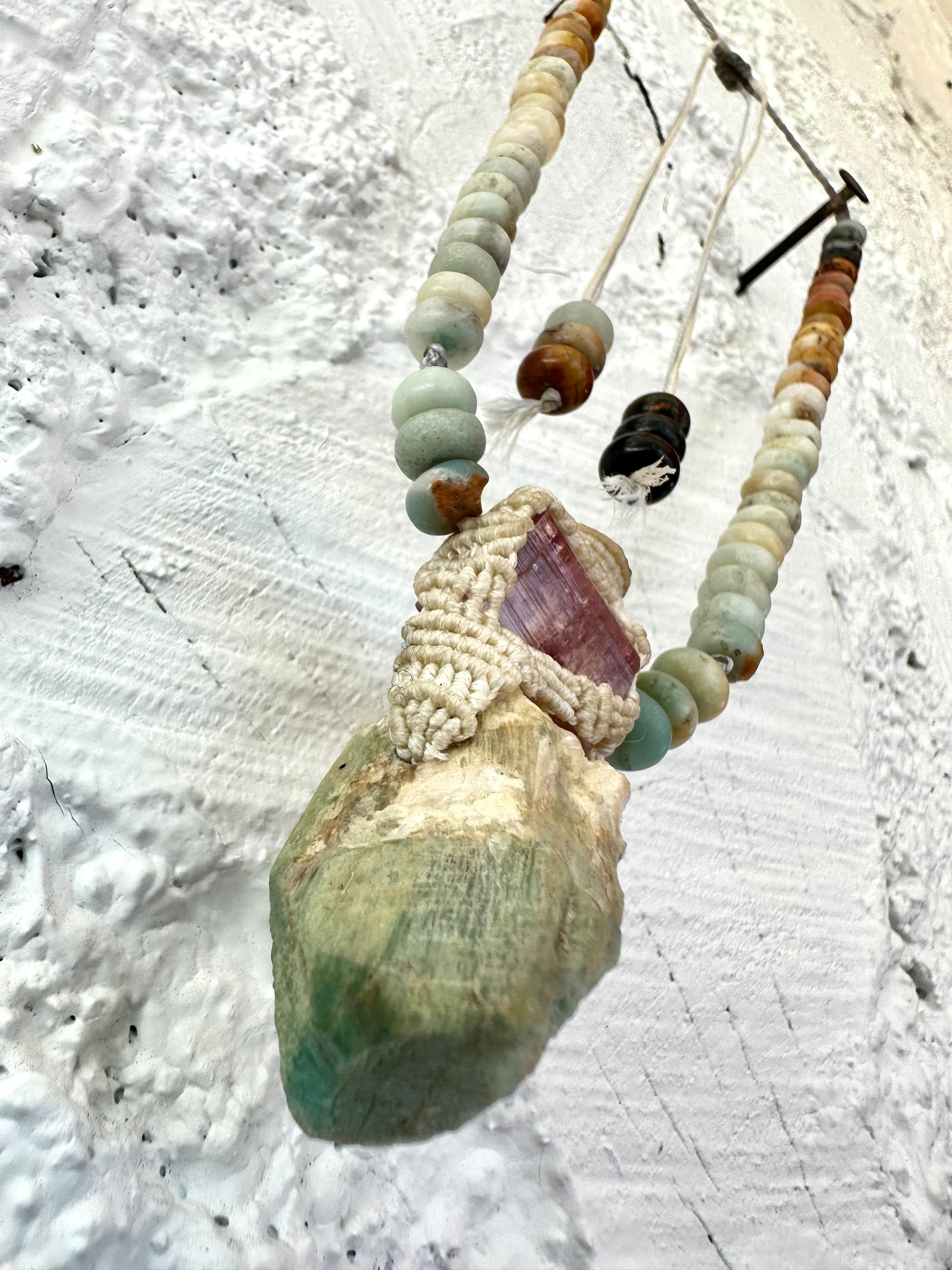 Detail of A luminous, pink tourmaline, soft turquoise amazonite stone And knotwork Basket necklace With beads of grated blue to white to brown amazonite Against a white painted cordwood wall