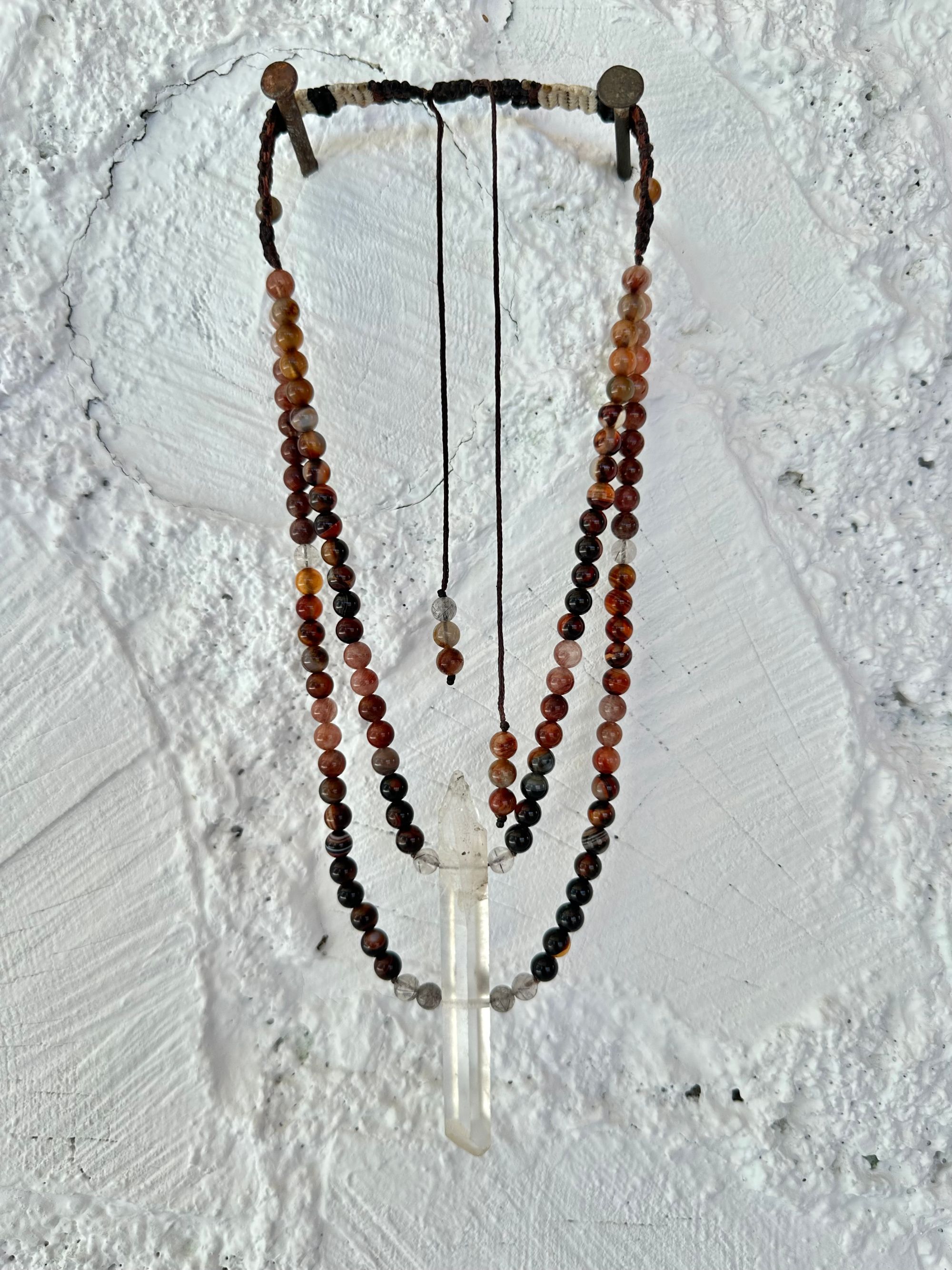 A necklace of clear quartz point with beads of deep hues of red ocre and soft pink hang against a white cordwood wall