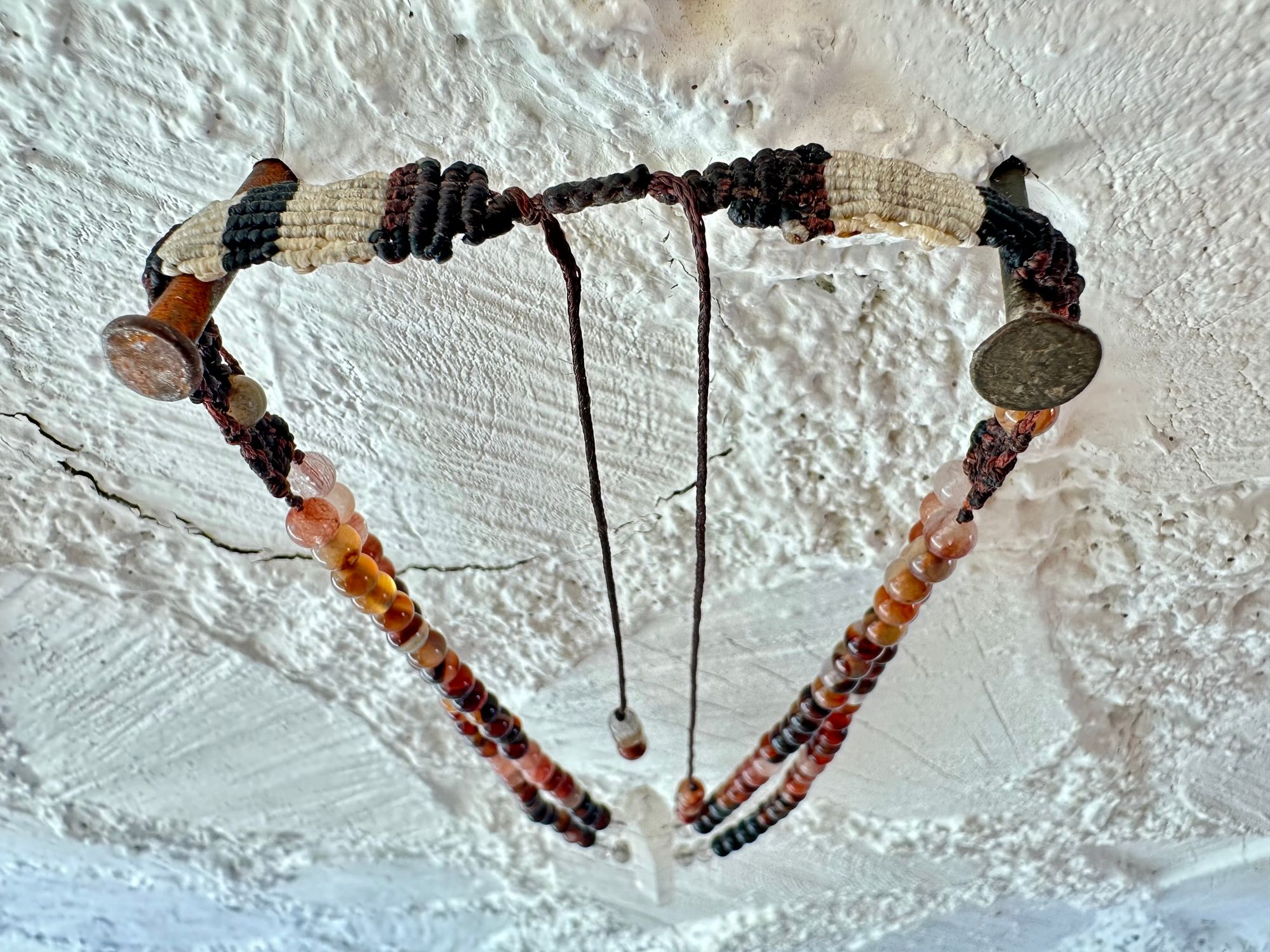 A necklace of clear quartz point with beads of deep hues of red ocre and soft pink hang against a white cordwood wall