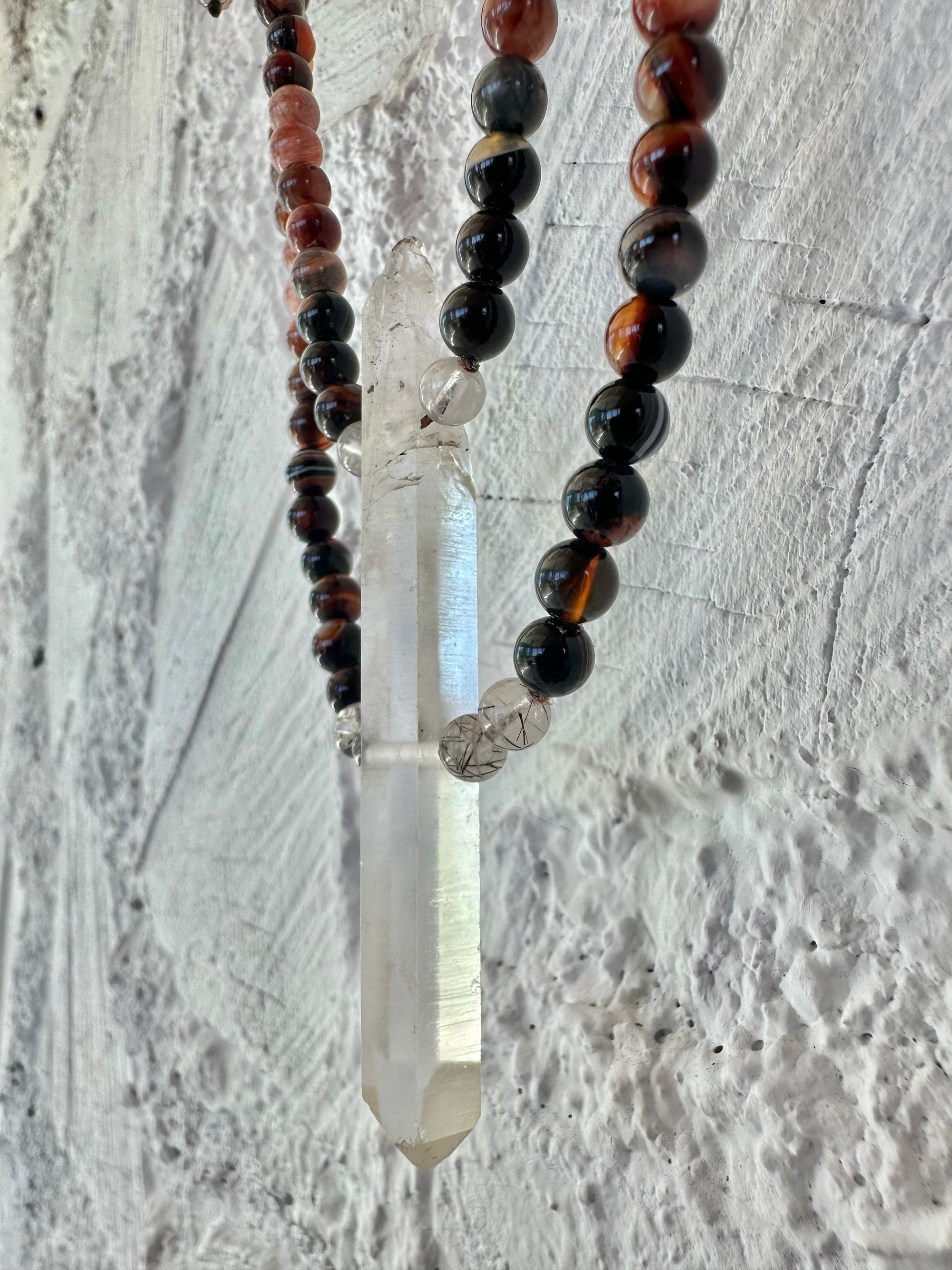 A necklace of clear quartz point with beads of deep hues of red ocre and soft pink hang against a white cordwood wall