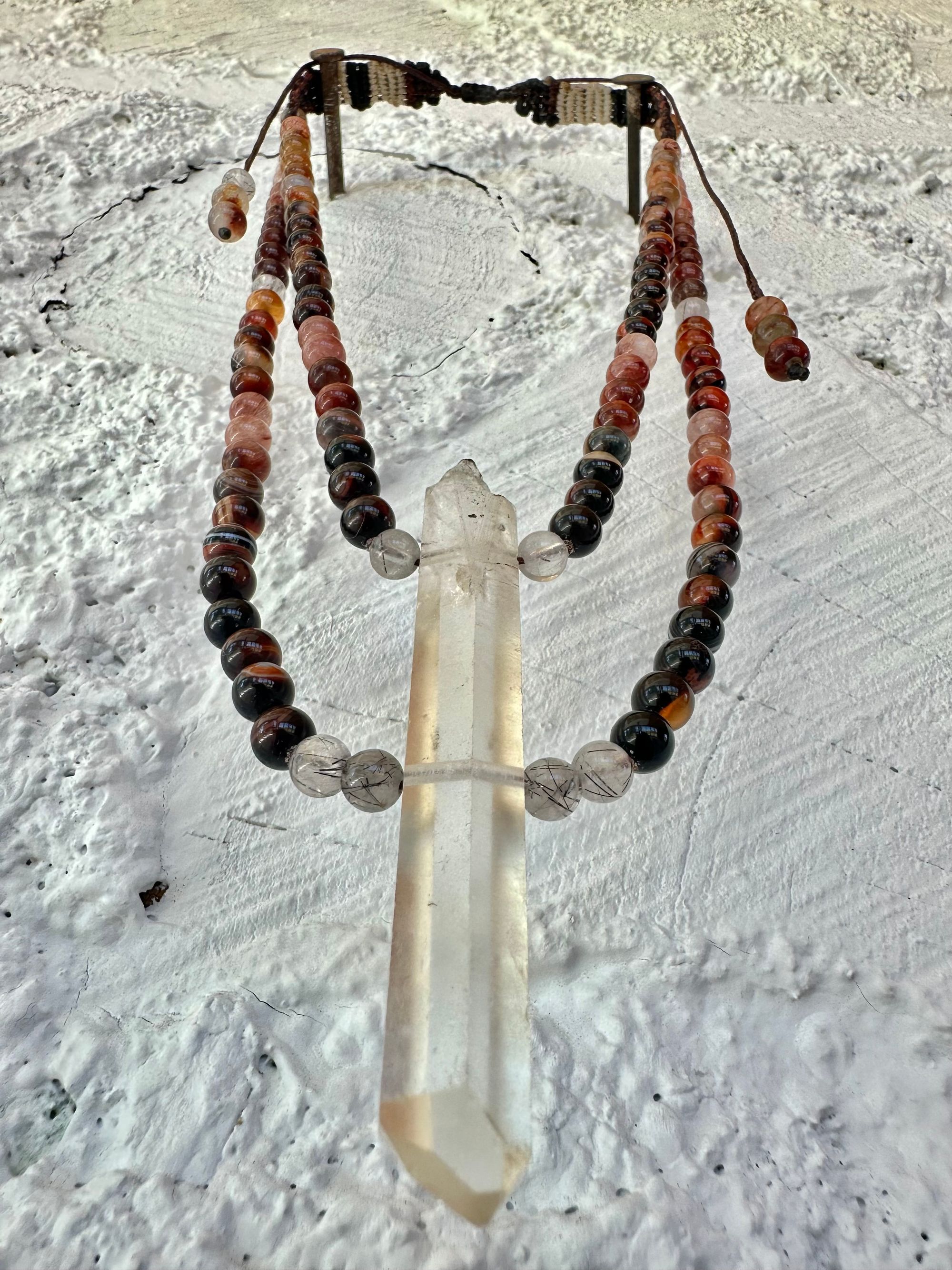 A necklace of clear quartz point with beads of deep hues of red ocre and soft pink hang against a white cordwood wall