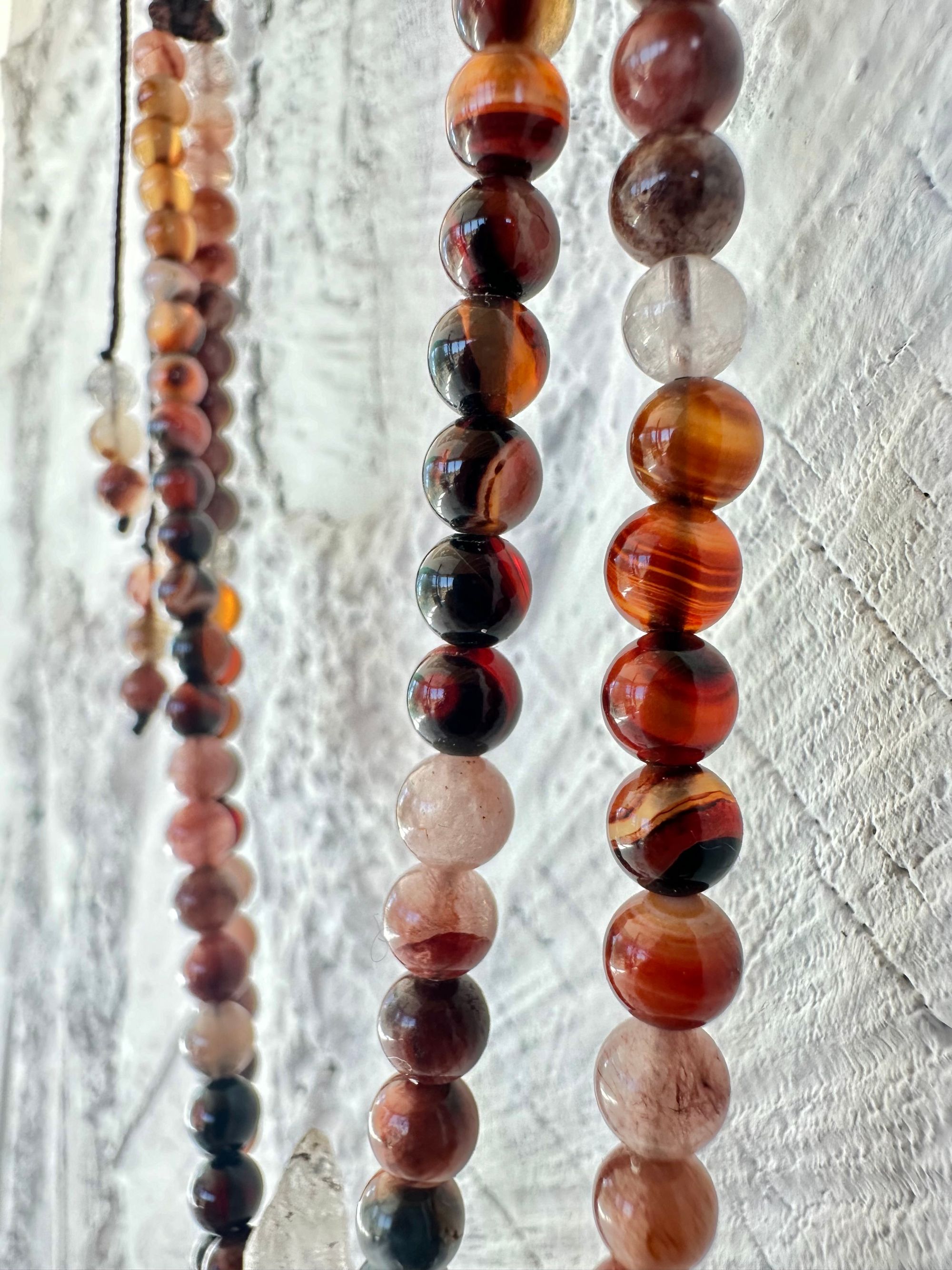 A necklace of clear quartz point with beads of deep hues of red ocre and soft pink hang against a white cordwood wall