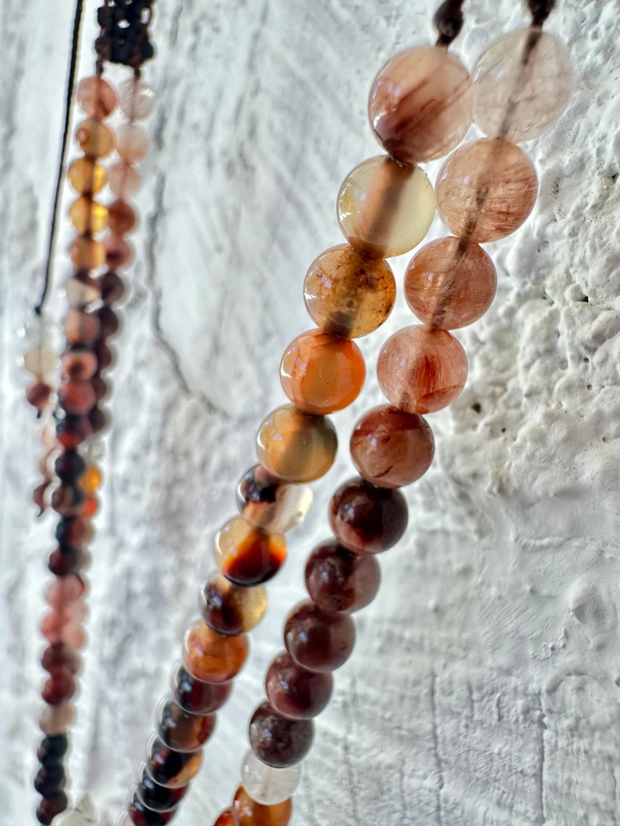 A necklace of clear quartz point with beads of deep hues of red ocre and soft pink hang against a white cordwood wall