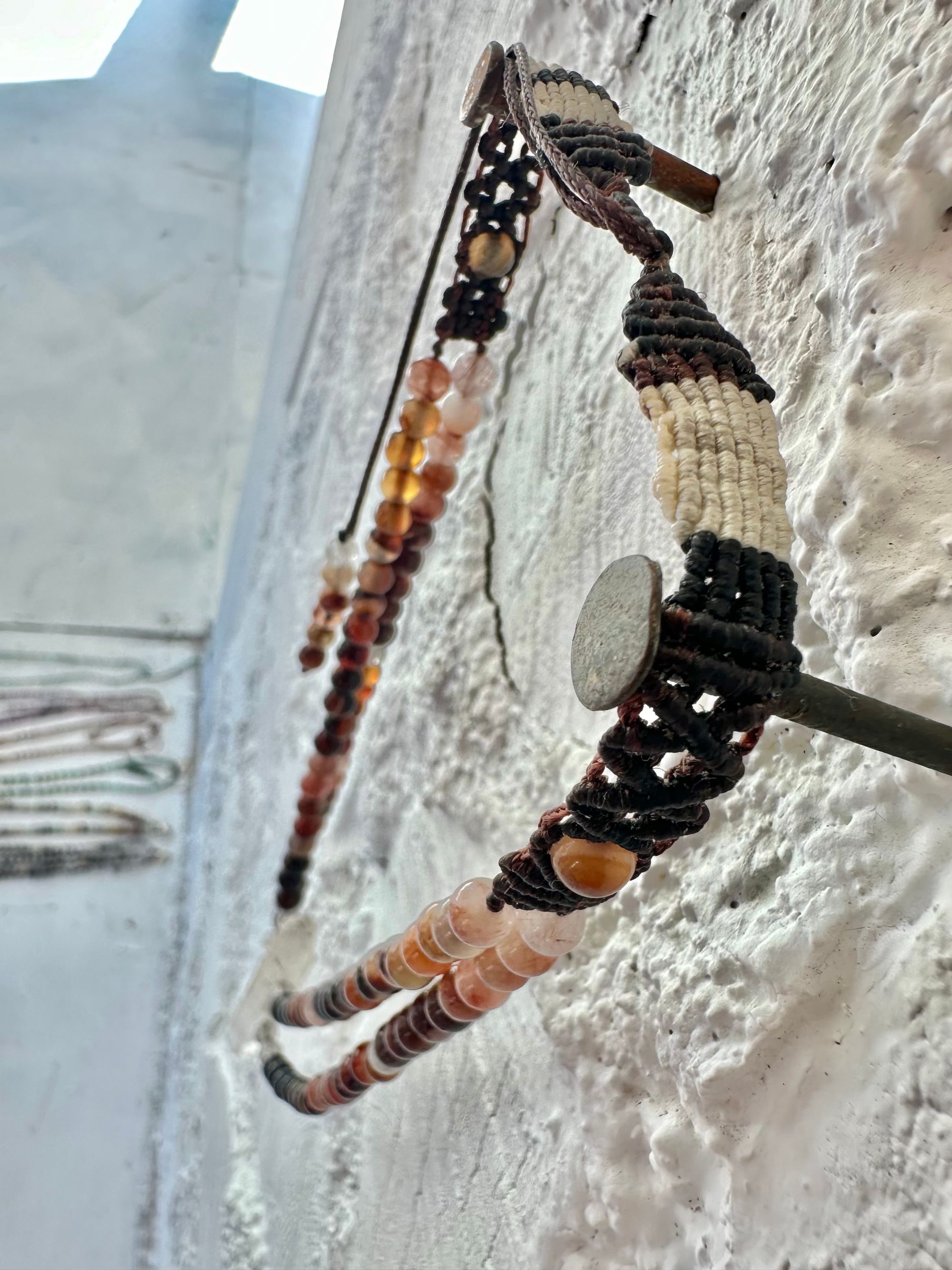 A necklace of clear quartz point with beads of deep hues of red ocre and soft pink hang against a white cordwood wall