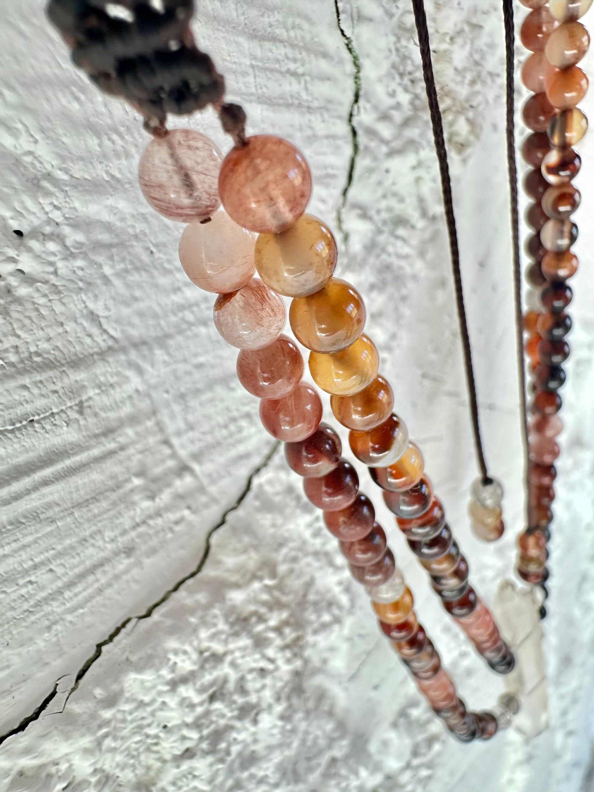 A necklace of clear quartz point with beads of deep hues of red ocre and soft pink hang against a white cordwood wall