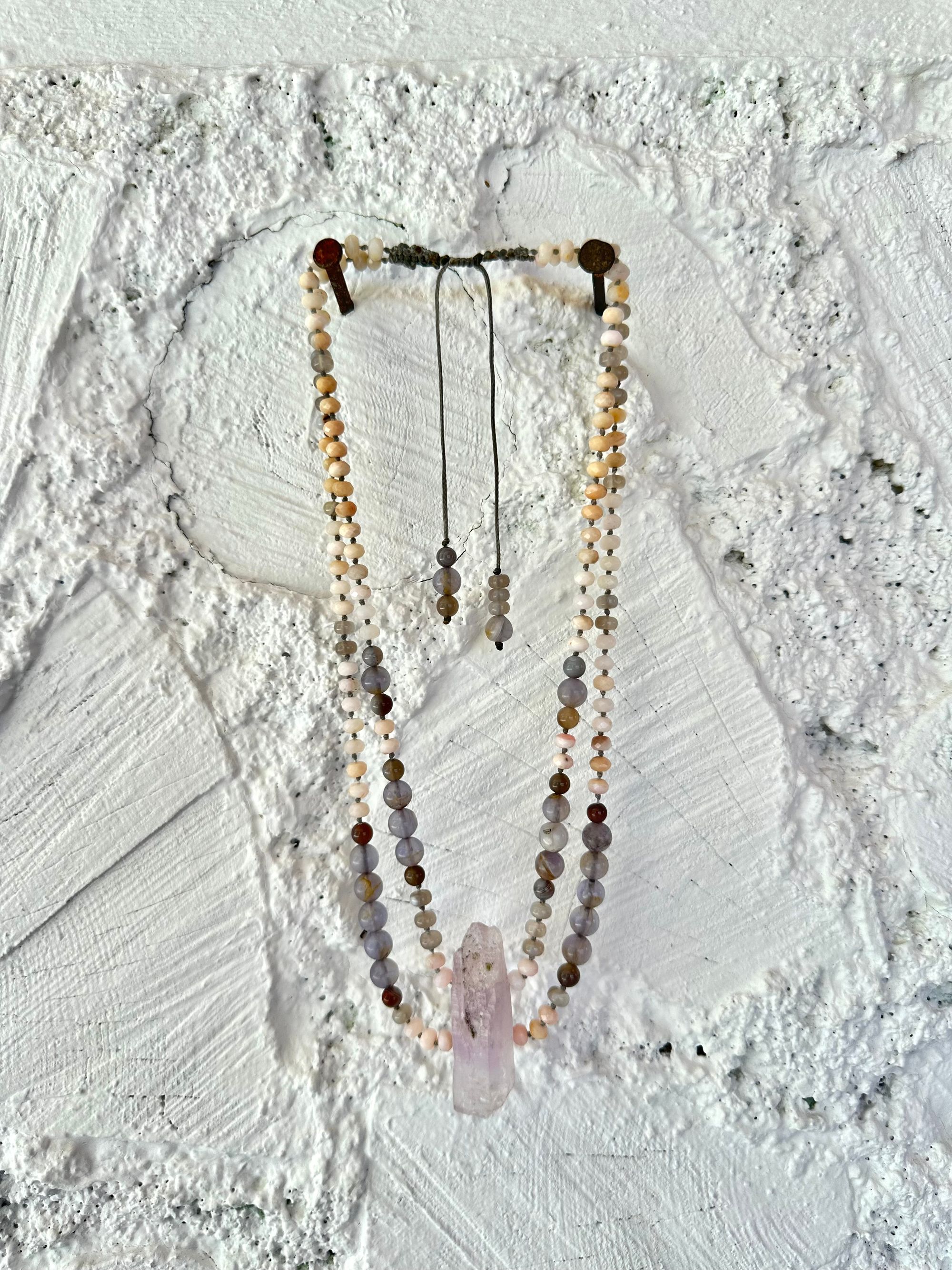 A necklace of soft but luminous, pink, purple and blue crystals hangs on a wall made of white painted cordwood