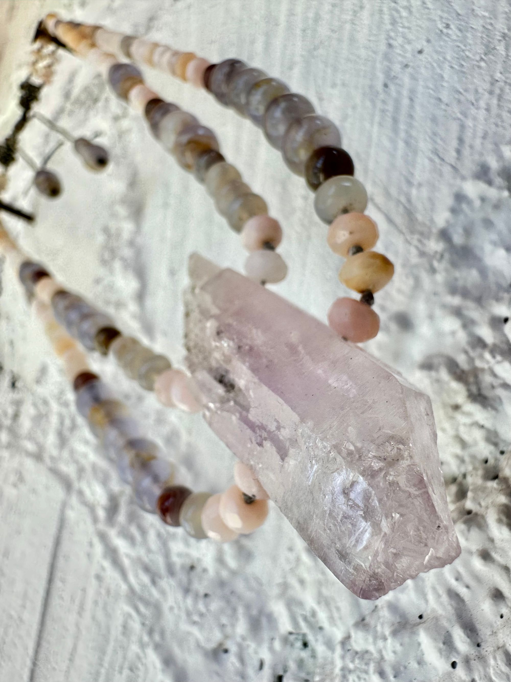 A necklace of soft but luminous, pink, purple and blue crystals hangs on a wall made of white painted cordwood