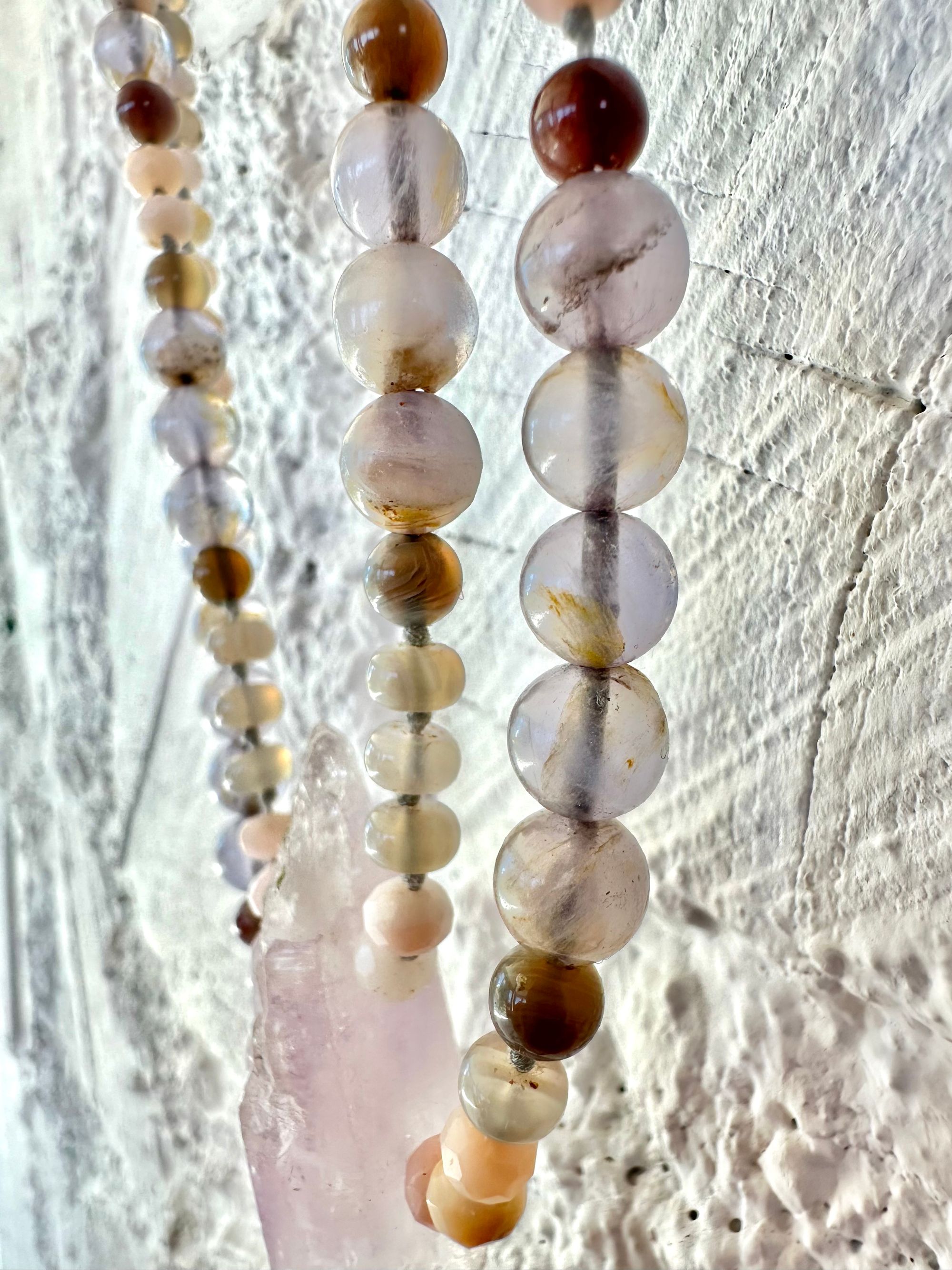 A necklace of soft but luminous, pink, purple and blue crystals hangs on a wall made of white painted cordwood