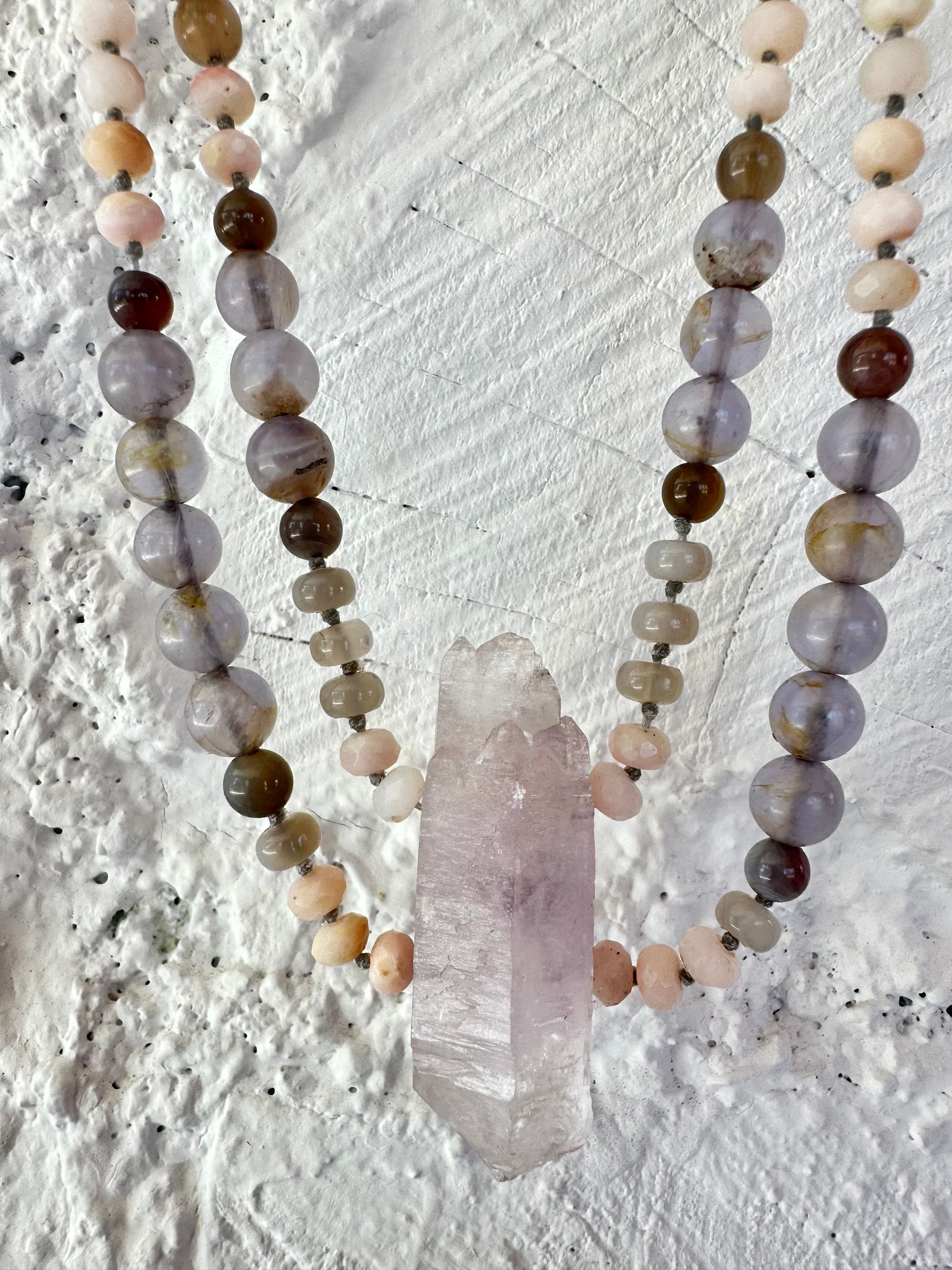 A necklace of soft but luminous, pink, purple and blue crystals hangs on a wall made of white painted cordwood