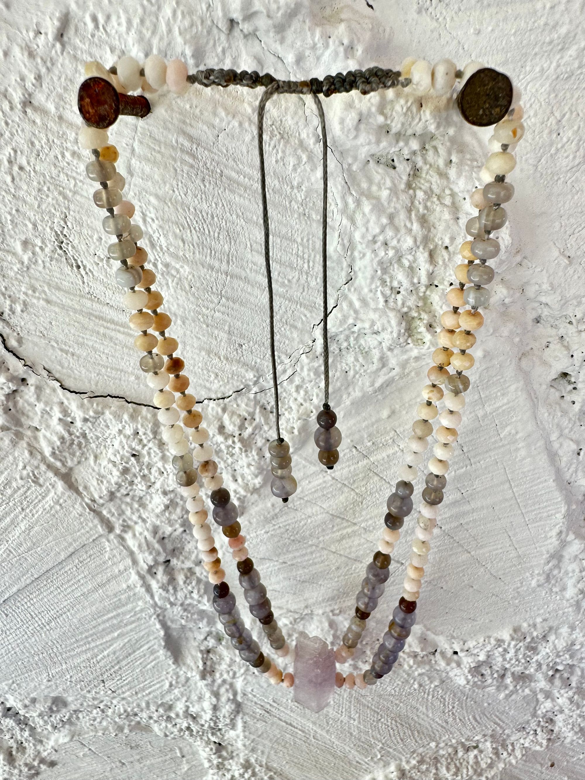 A necklace of soft but luminous, pink, purple and blue crystals hangs on a wall made of white painted cordwood